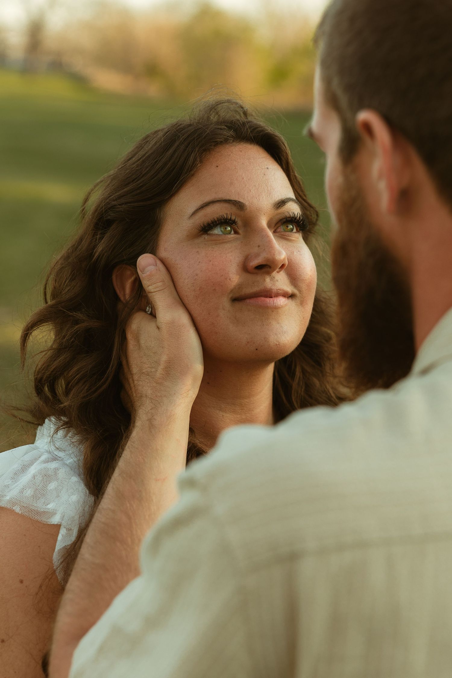 A bearded man tenderly cups a smiling brunette woman's face outdoors during golden hour.