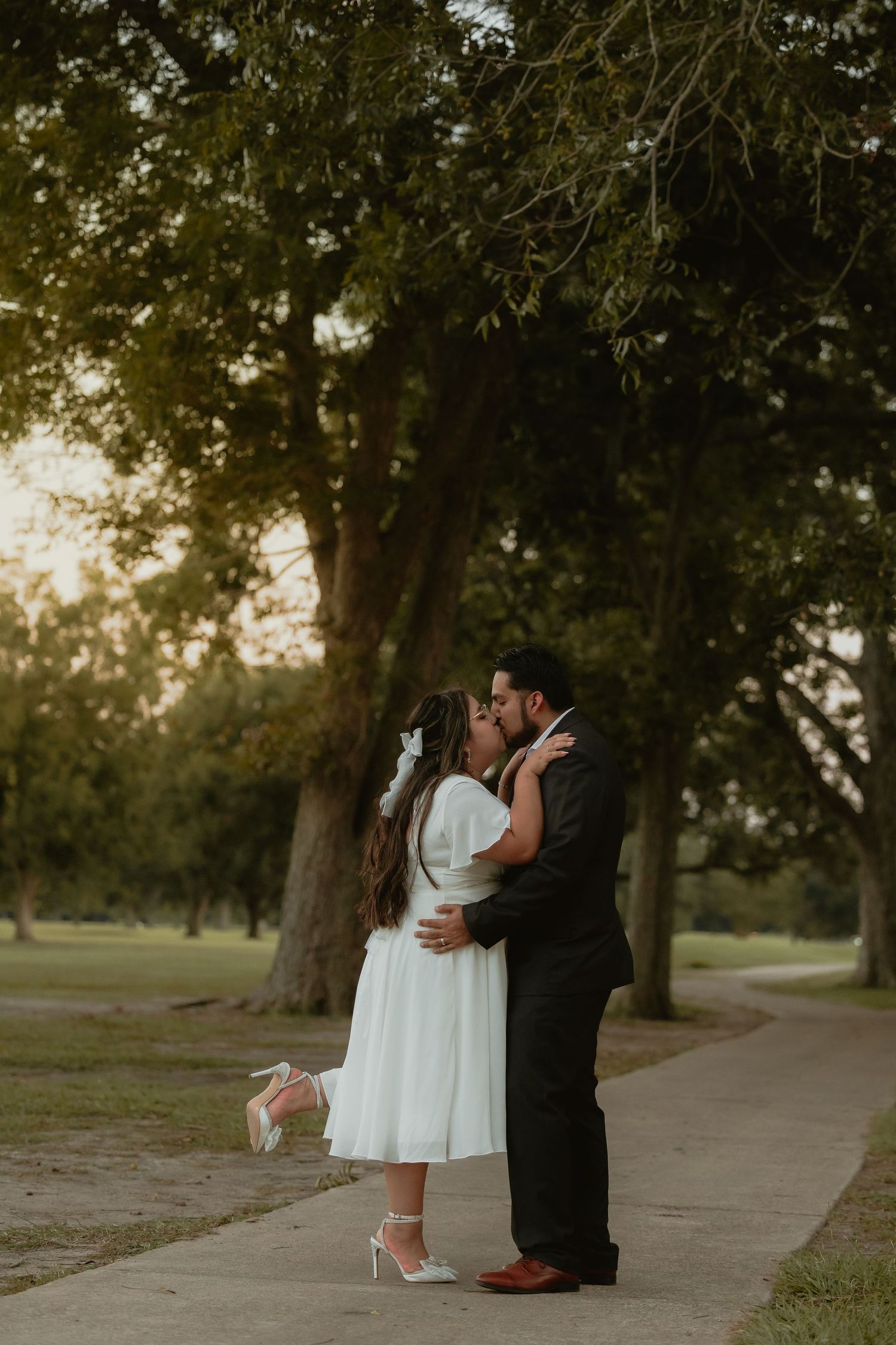 Intimate moment between couple in formal wear on path beneath tall trees.