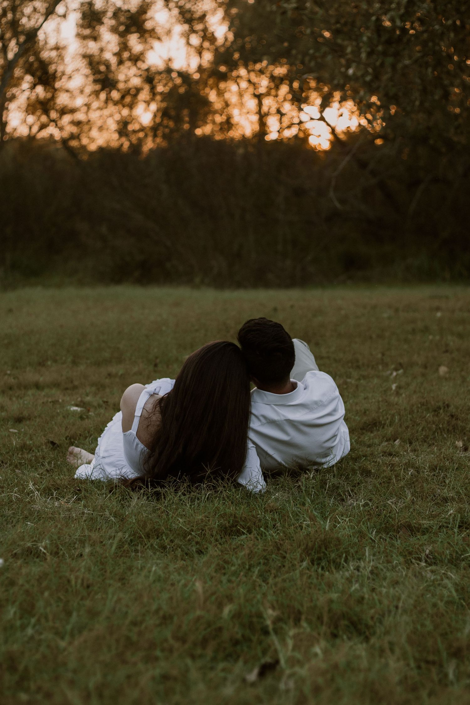 Two people in white clothing lie close together on grass during a moody sunset photoshoot.