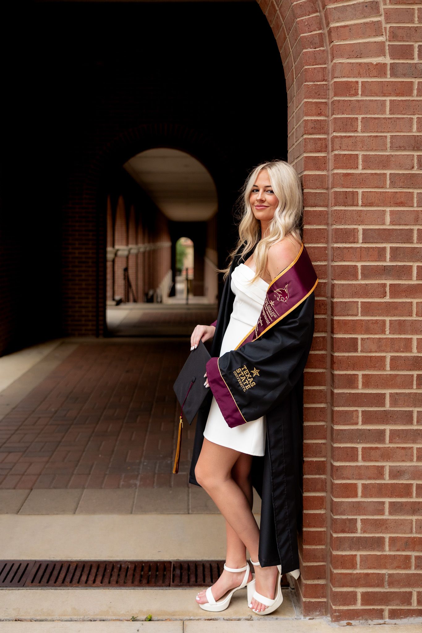 Blonde woman in white dress, black graduation gown and maroon sash poses by brick arch colonnade.