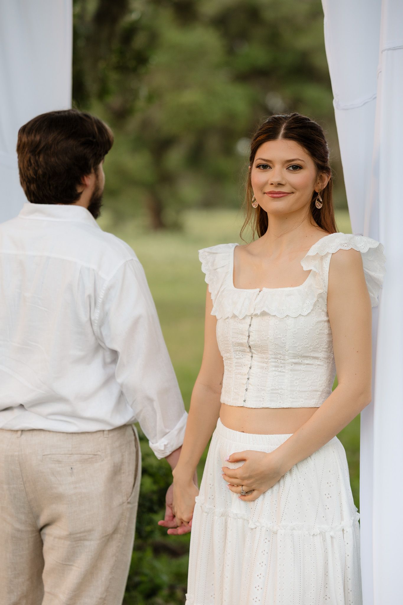 A bride in white crop top and skirt holds hands with groom in white shirt at an outdoor wedding ceremony.