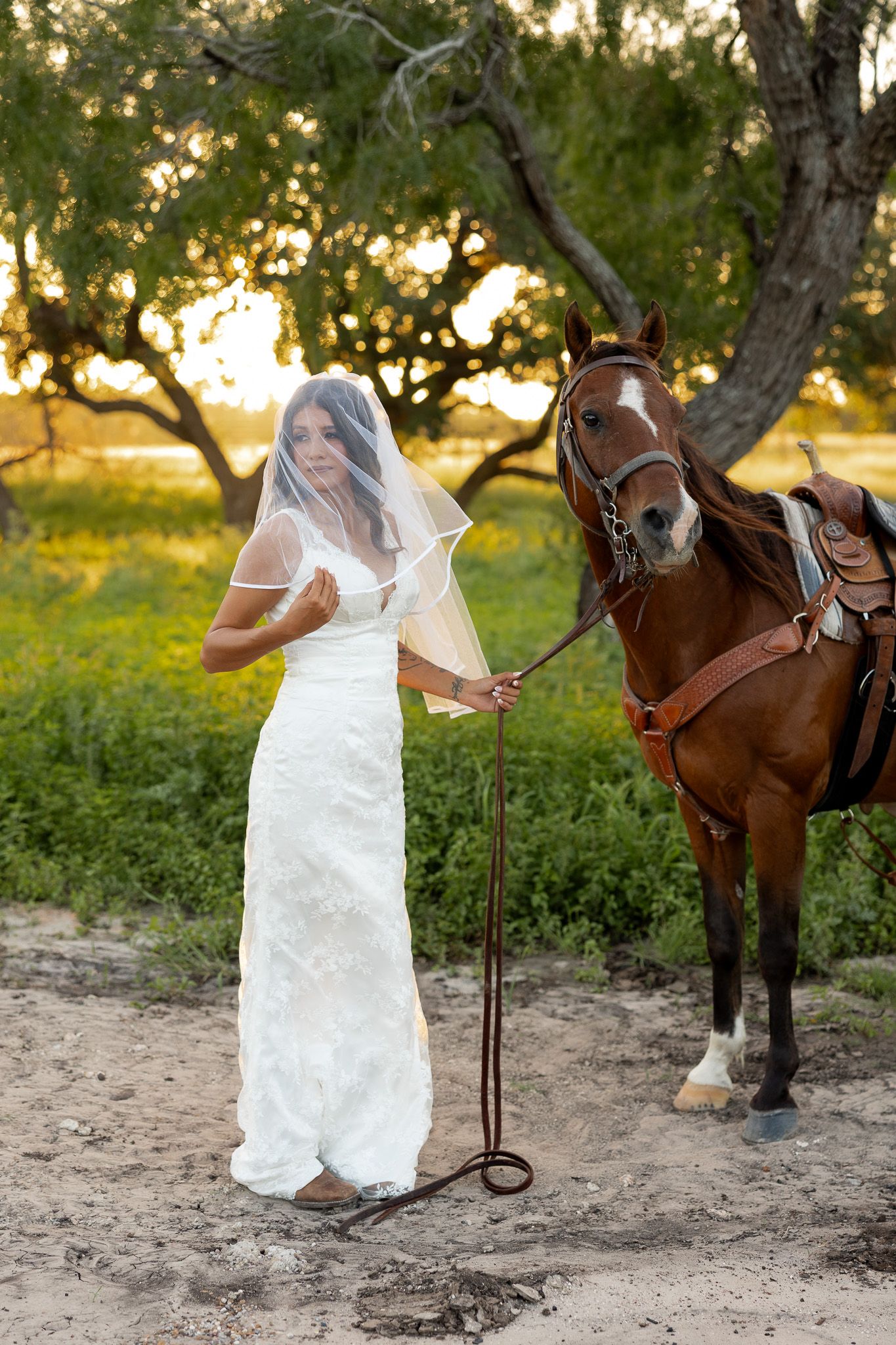 Bride in white lace gown and veil stands beside a brown horse at sunset in a scenic outdoor setting.