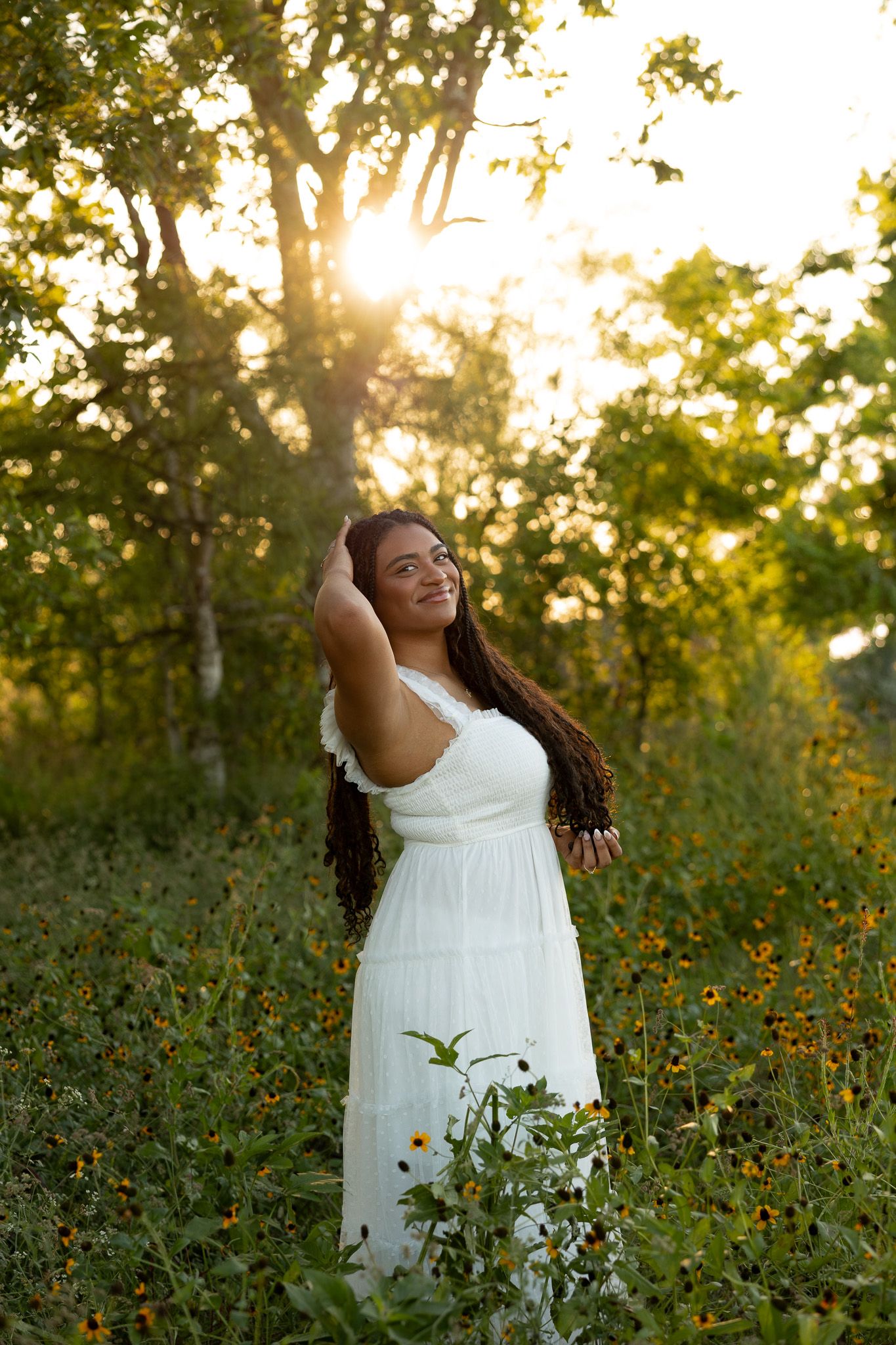 Young woman in white dress standing in wildflower field at golden hour sunset with trees in background.