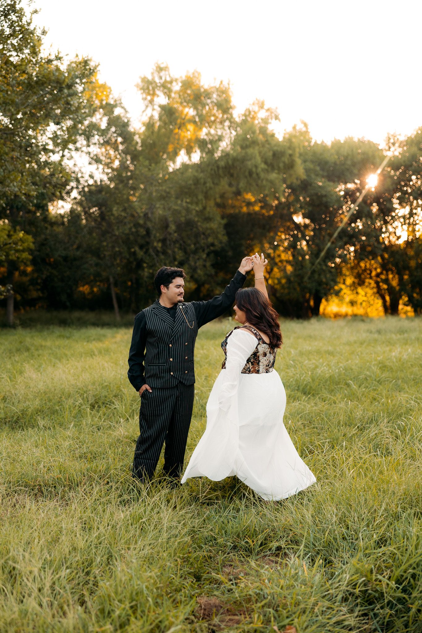 Couple dancing in golden hour sunlit meadow, woman in white gown twirling, man in black suit, lush green trees background.
