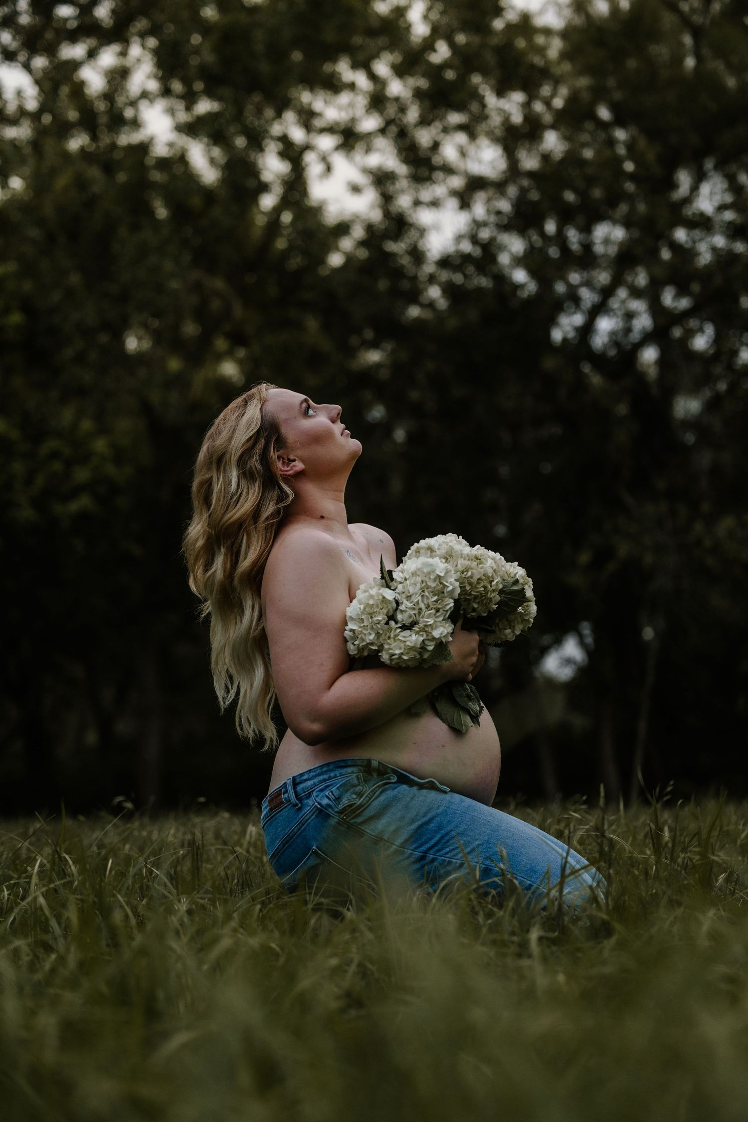 A candid maternity photo shows a woman holding baby's breath flowers while sitting in tall grass.