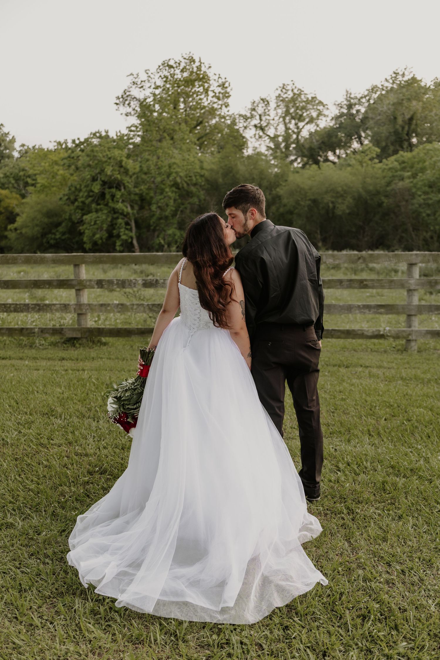 Couple in wedding attire dancing in a rustic outdoor setting with wooden fence.