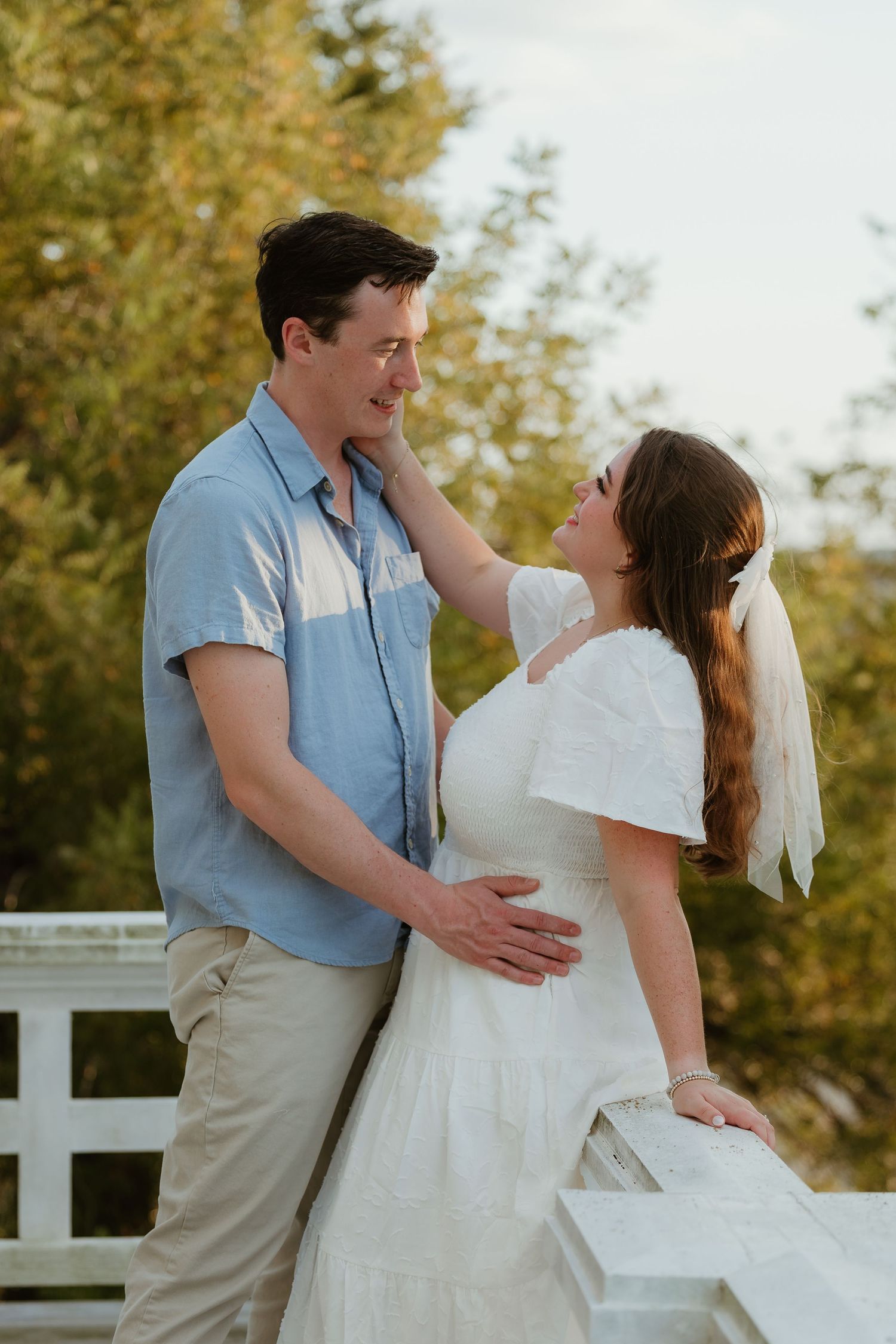 A couple in white and blue outfits embraces near autumn trees.