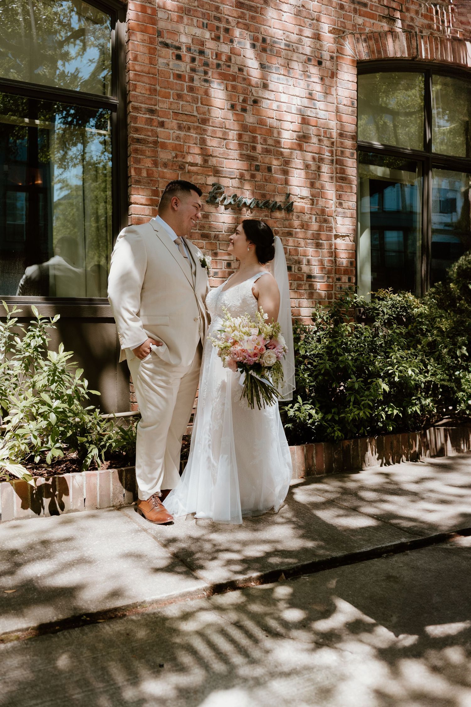 Sunlit wedding portraits against rustic brick wall with lush greenery.