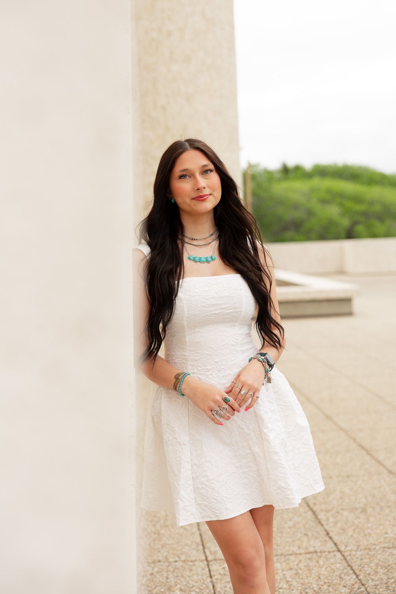 Young woman in white dress with turquoise jewelry leans against marble column outdoors.