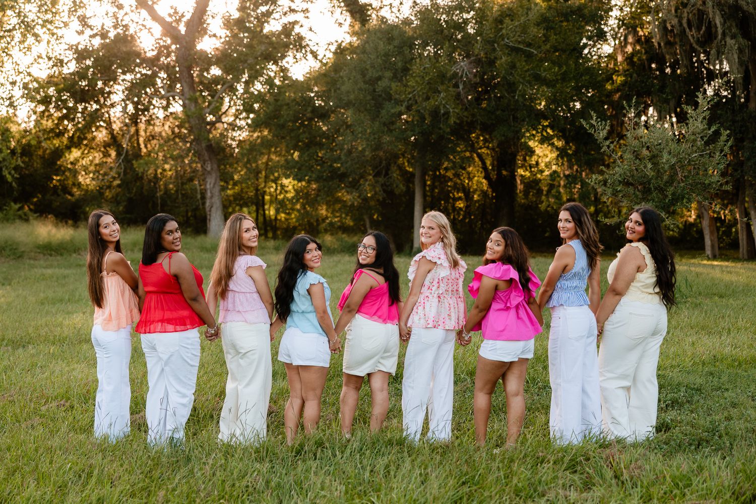 Group of eight women wearing vibrant tops and white pants smiling outdoors in a lush green park setting.