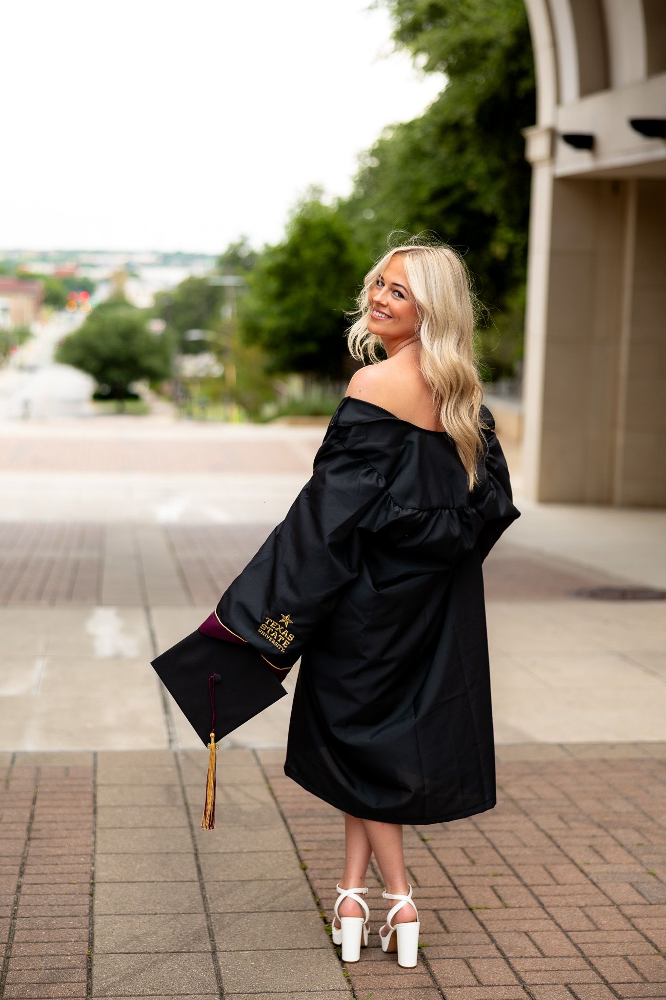 Smiling blonde woman in black graduation gown and cap posing outdoors on campus in white heels.