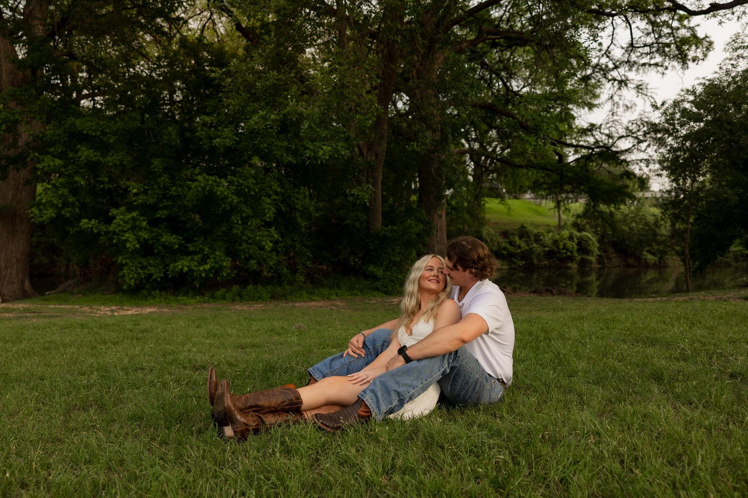 Couple sitting together on lush green grass, leaning close romantically, surrounded by tall trees in soft natural light.