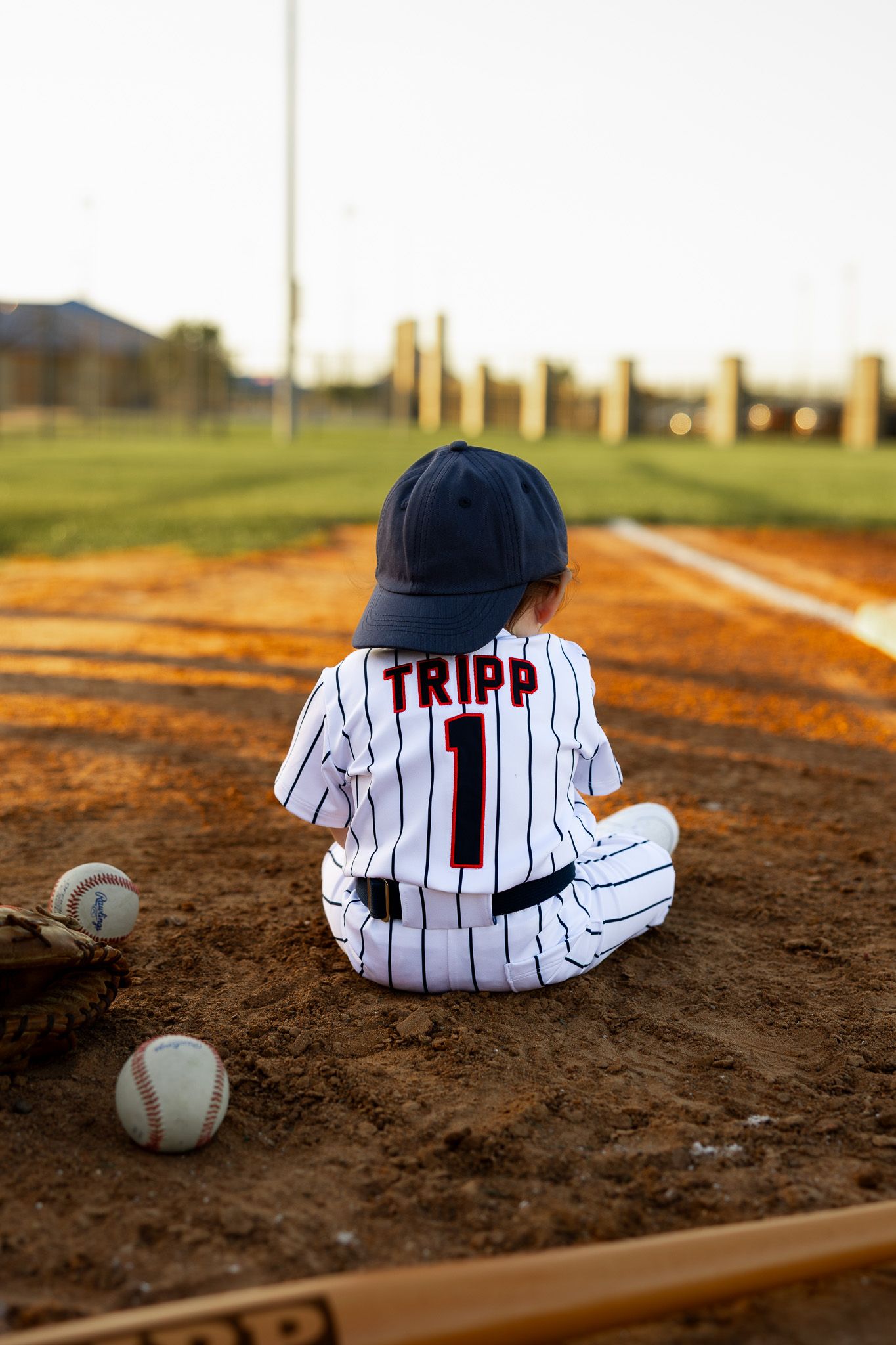 Toddler in Tripp #1 pinstripe baseball uniform sitting on baseball diamond dirt with baseballs and glove.
