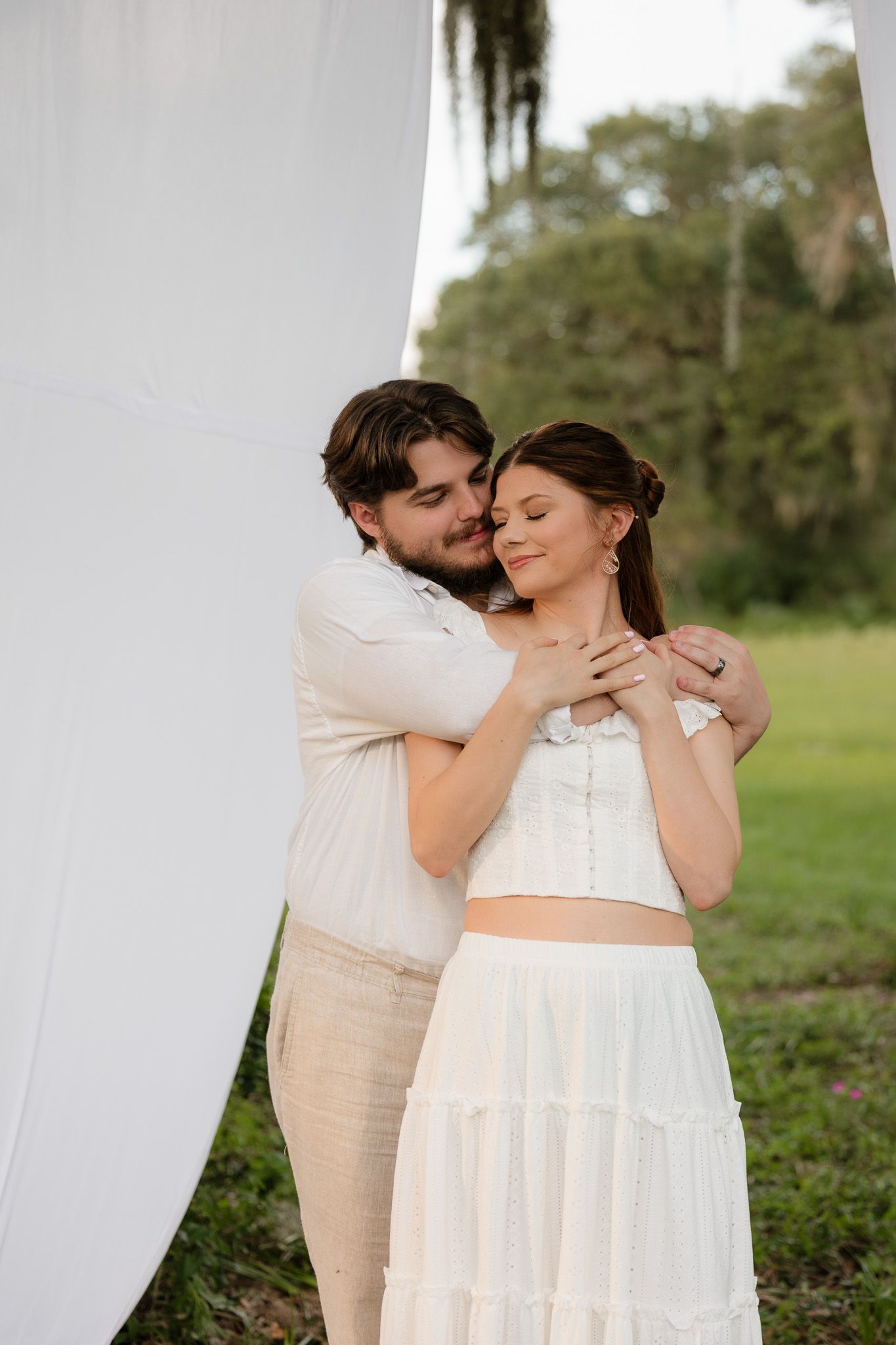 A couple in white attire embraces tenderly outdoors, surrounded by lush greenery and flowing white drapes.