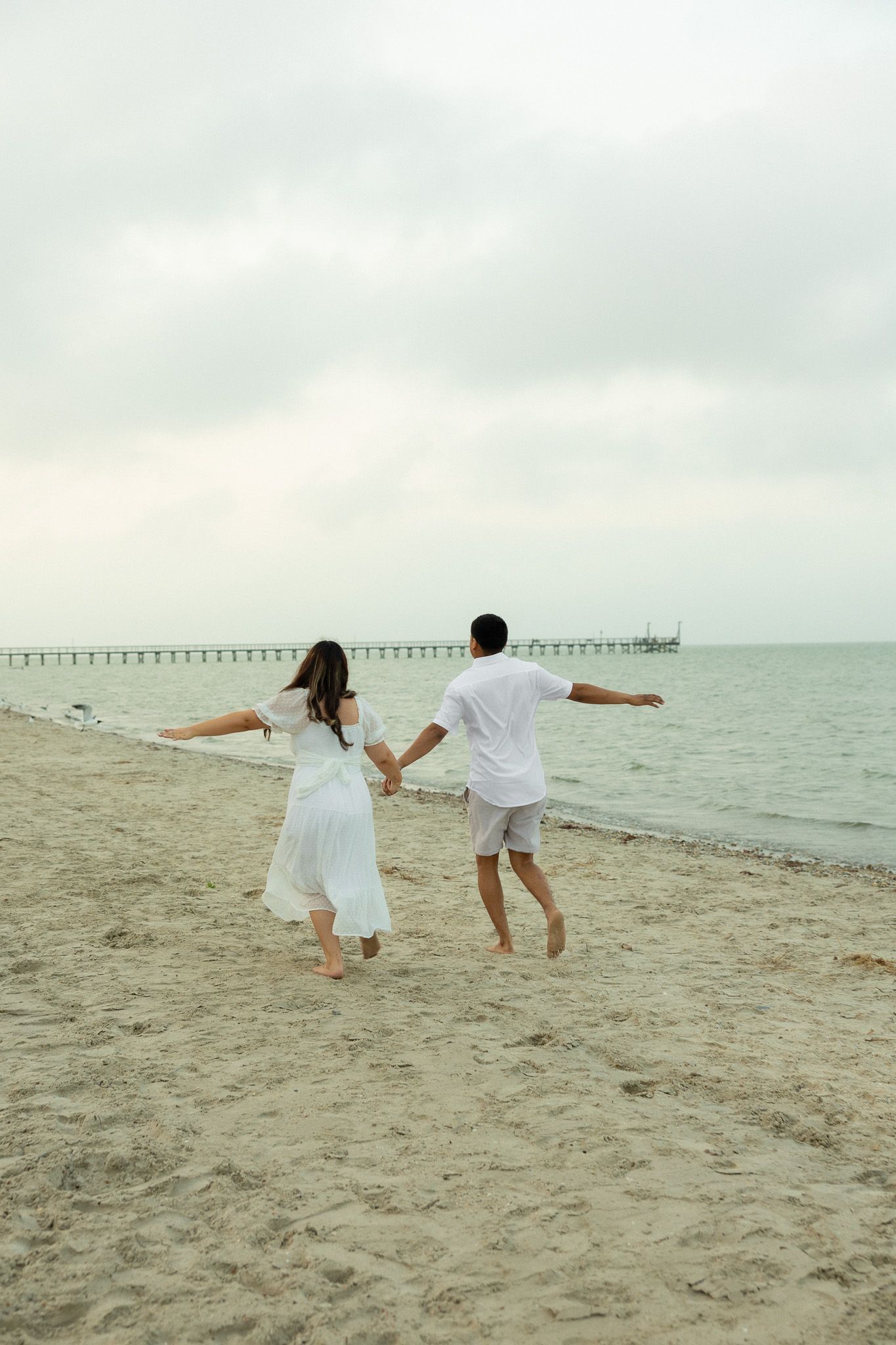 Couple in white outfits holding hands and running barefoot on a sandy beach with a pier in the background.