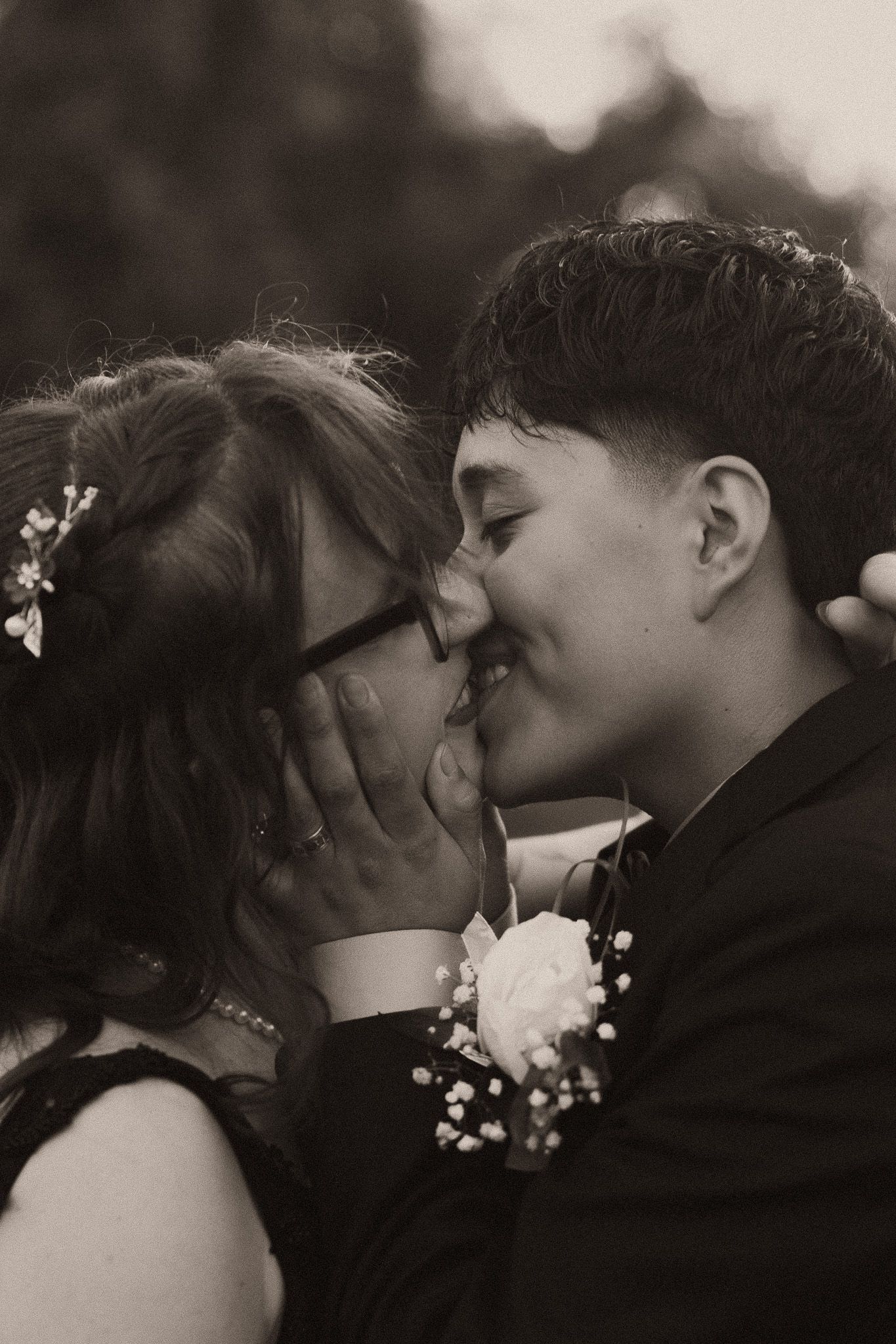 Black and white photo of a couple sharing a romantic kiss at prom, dressed in formal attire with corsage and hair flowers.
