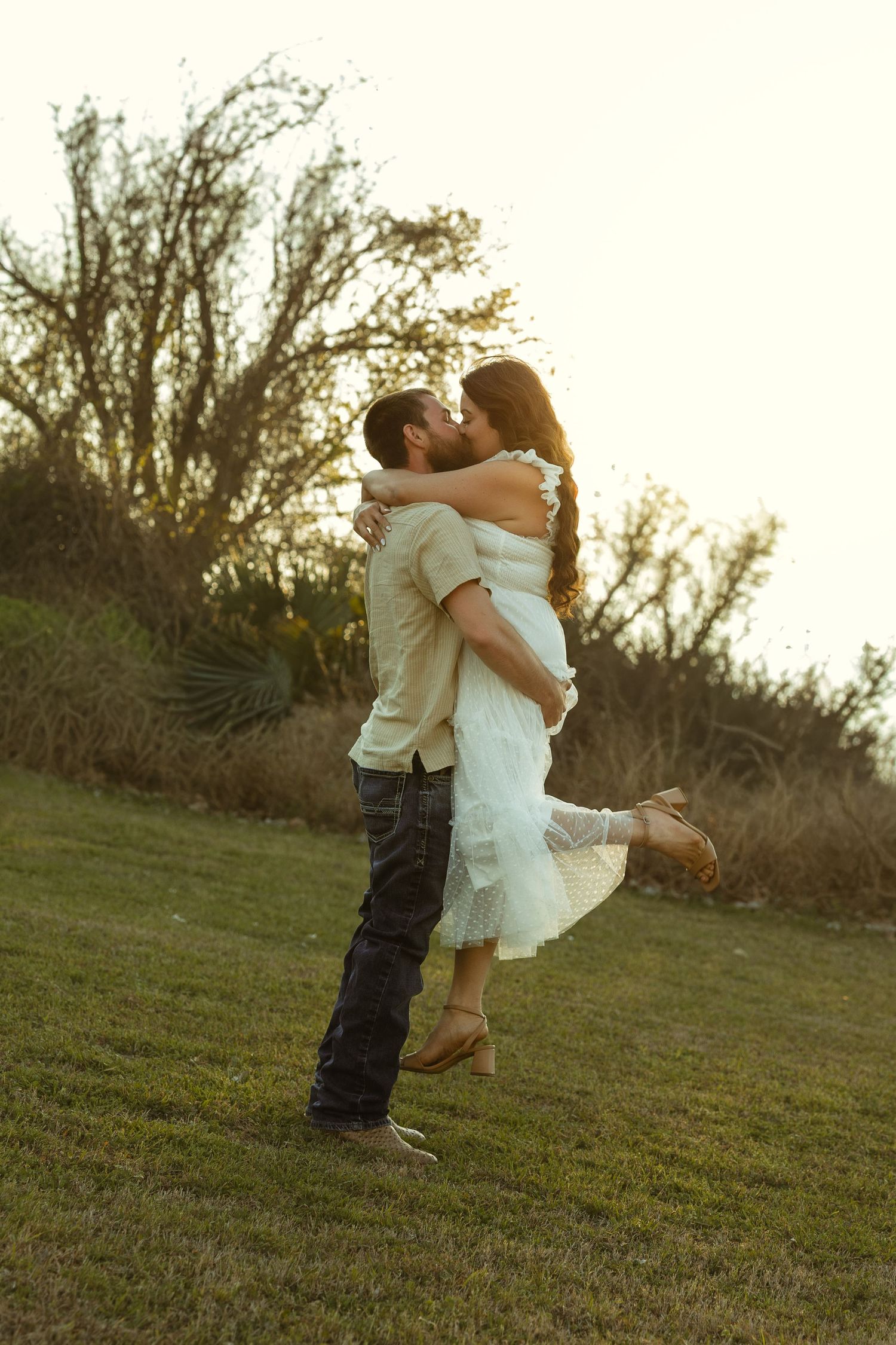 A couple shares a romantic kiss at sunset, man lifting woman in white dress on a grassy hill with trees behind.