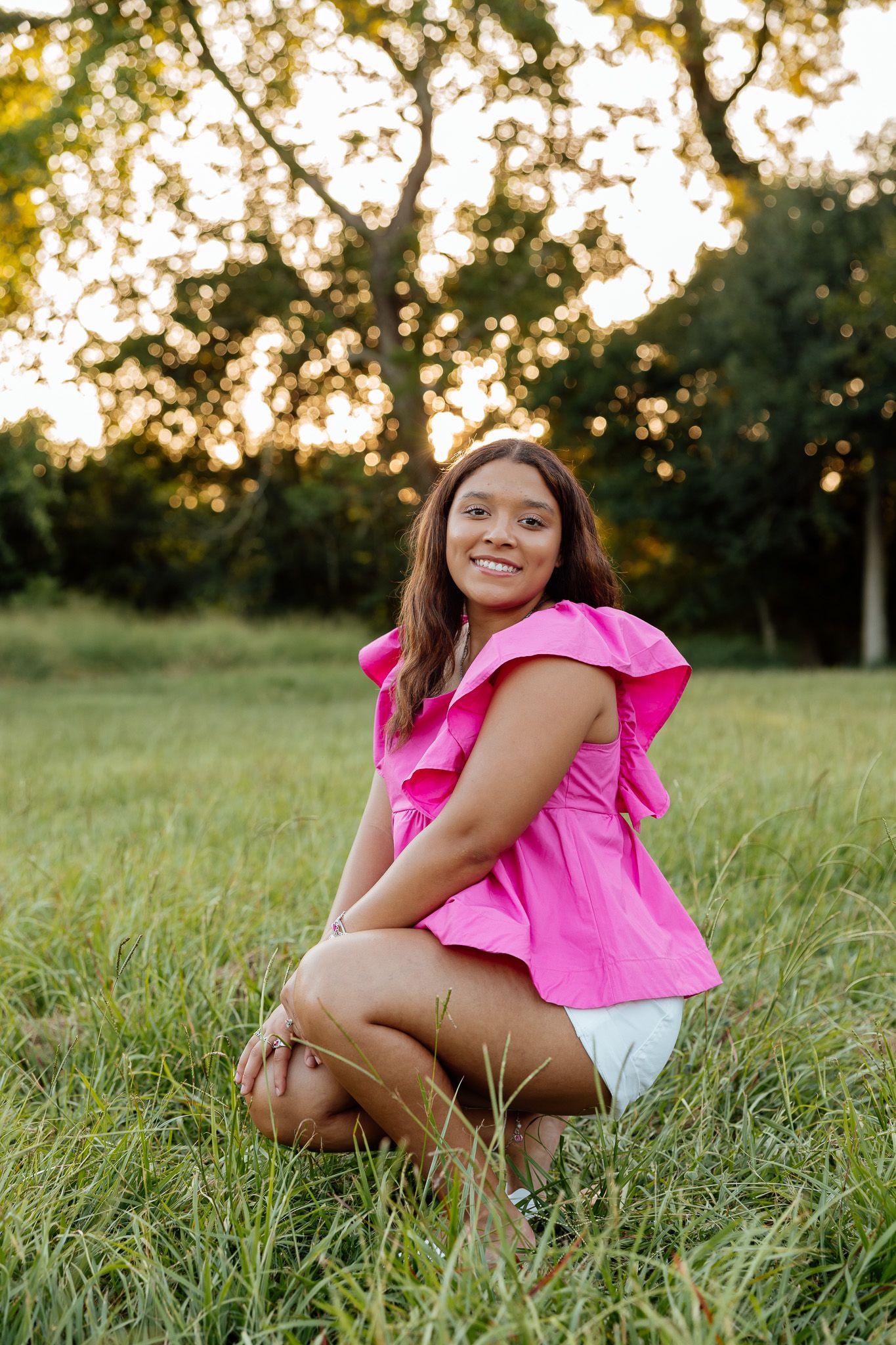 Young woman in pink ruffled top and white shorts smiling while kneeling in green grass at golden hour.