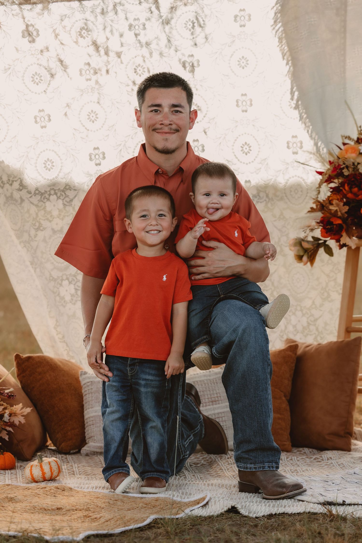 Father and two young sons in matching red shirts and jeans pose for a fall family portrait with pumpkins.