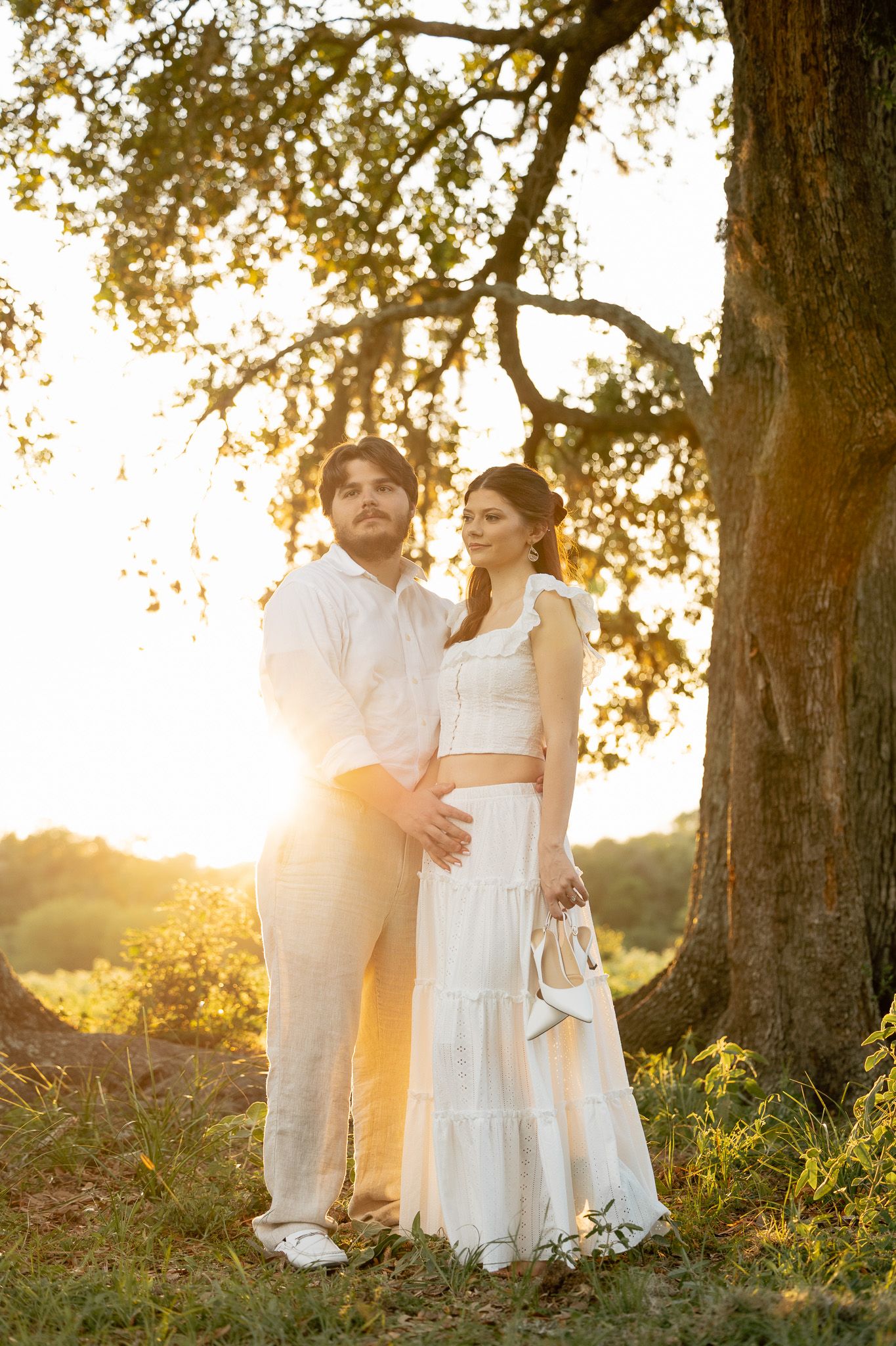Couple in white outfits embracing under a large tree during golden hour sunset in a serene outdoor setting.