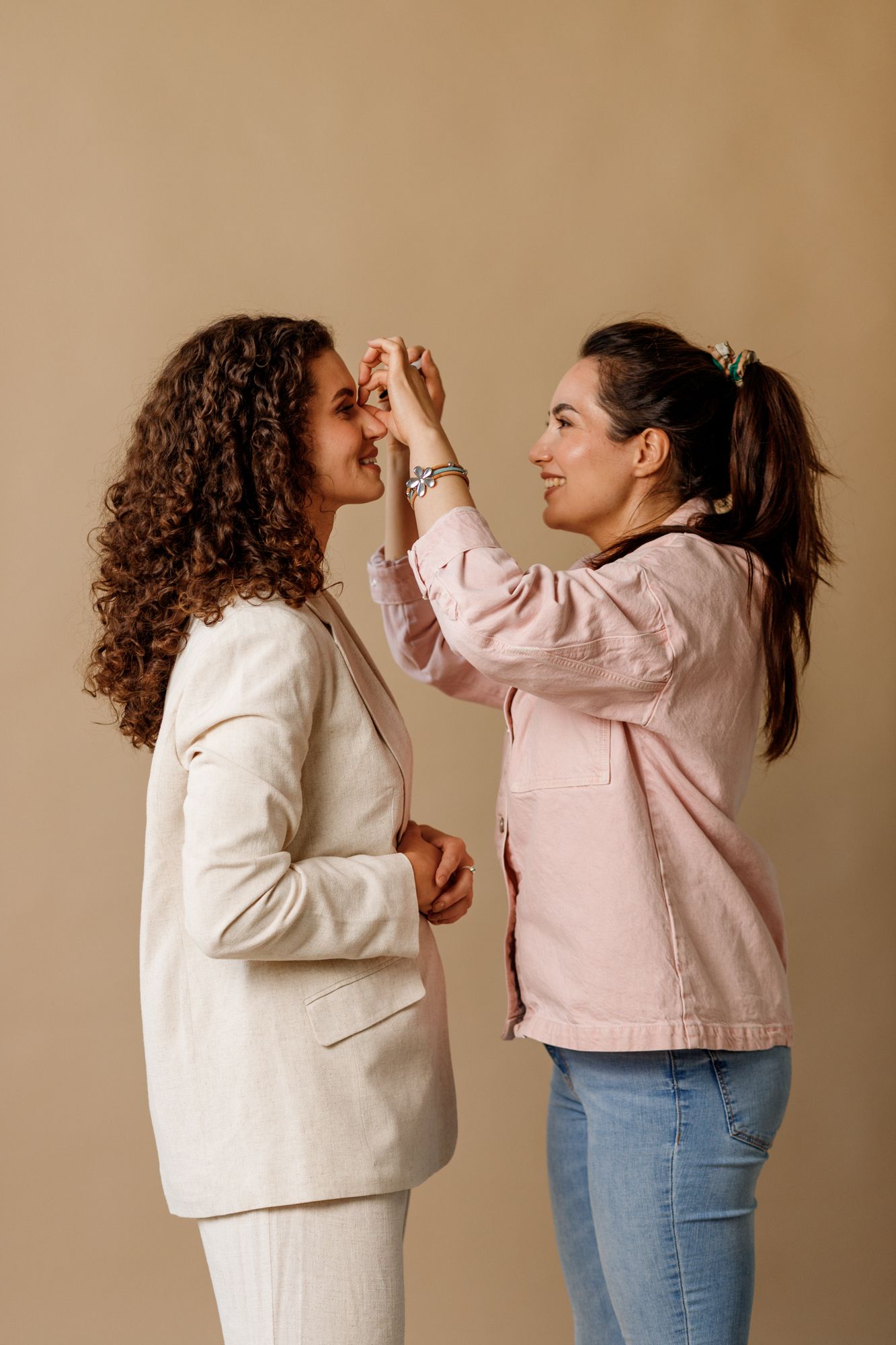Two stylish women in casual outfits interact playfully against a beige background as one person applies makeup to the other.