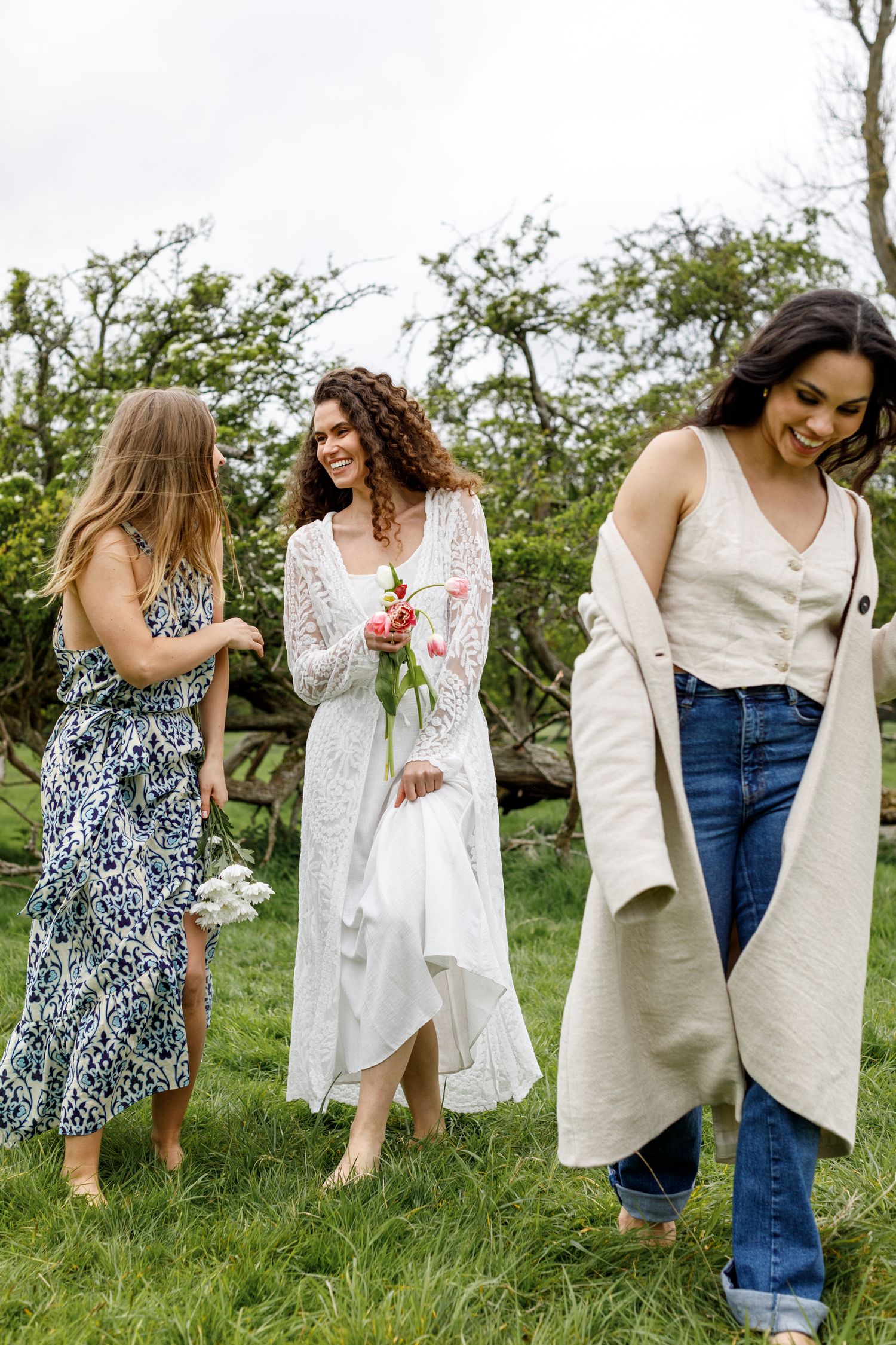 Three women in stylish spring outfits stand together on grassy hillside, one holding fresh flowers in a white dress.