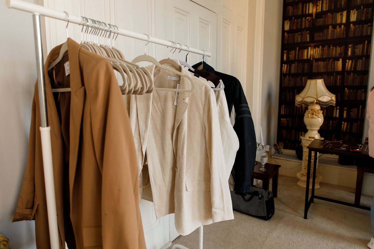 Minimalist clothing rack with neutral colored garments and table lamp near bookshelf in corner of room.