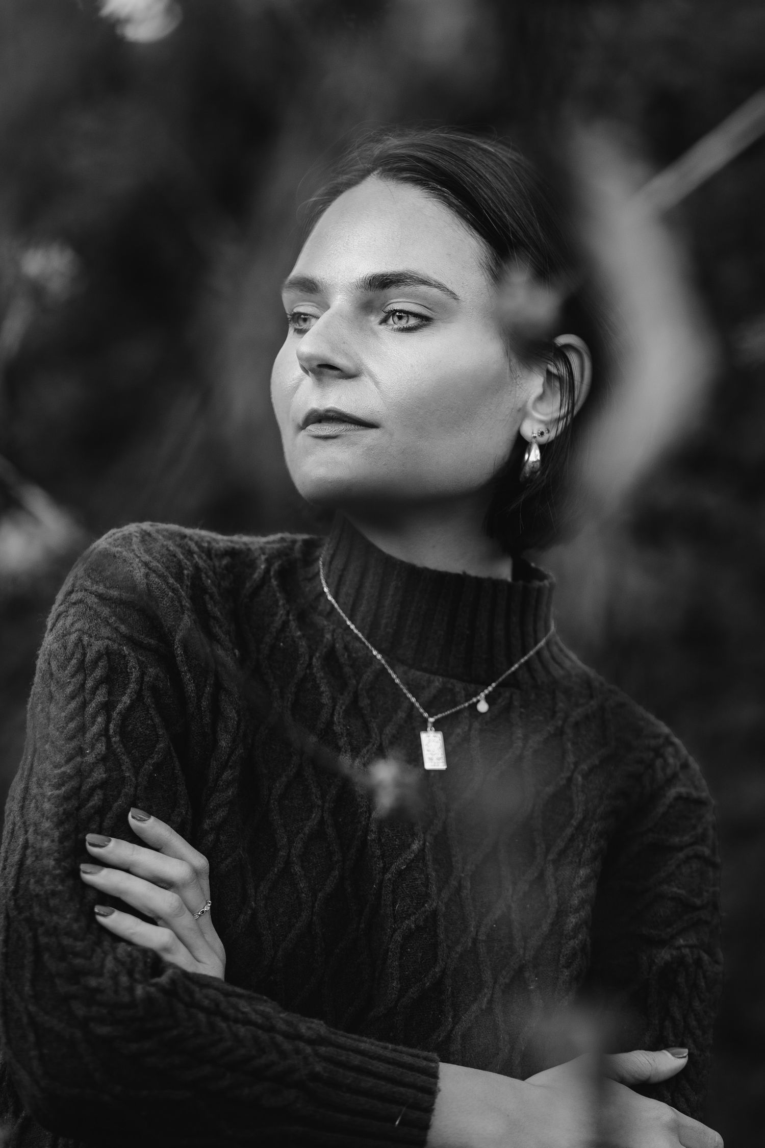 Black and white portrait of someone in a cable knit sweater wearing delicate jewelry against a blurred natural backdrop.