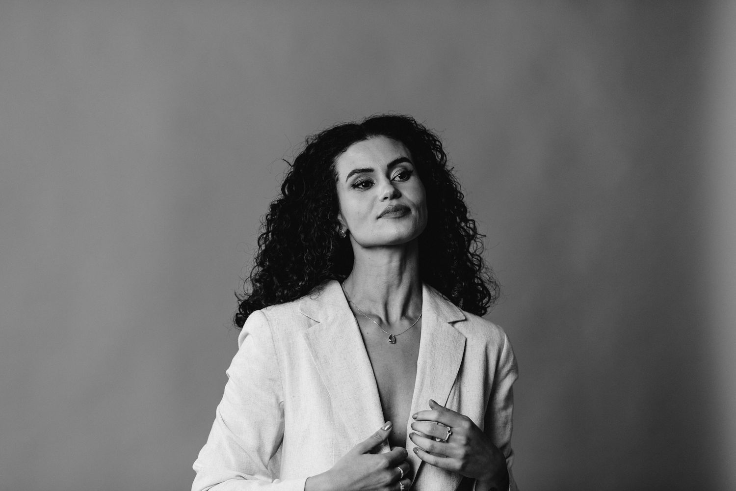 Black and white portrait of a person with curly hair wearing a white blazer against a simple studio backdrop.