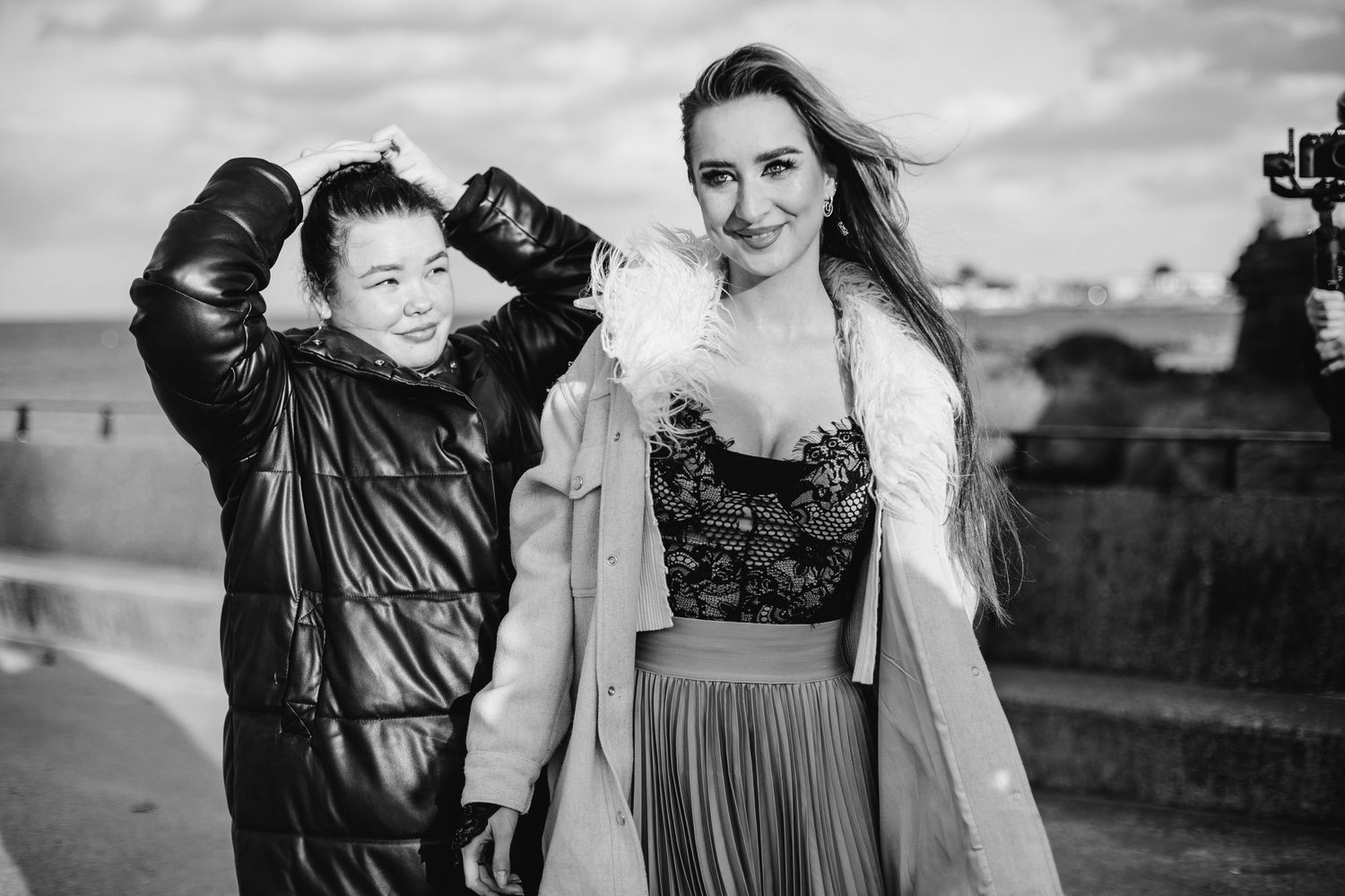 Two people pose together outdoors in black and white, one wearing a leather jacket and one in a dress with light outerwear.
