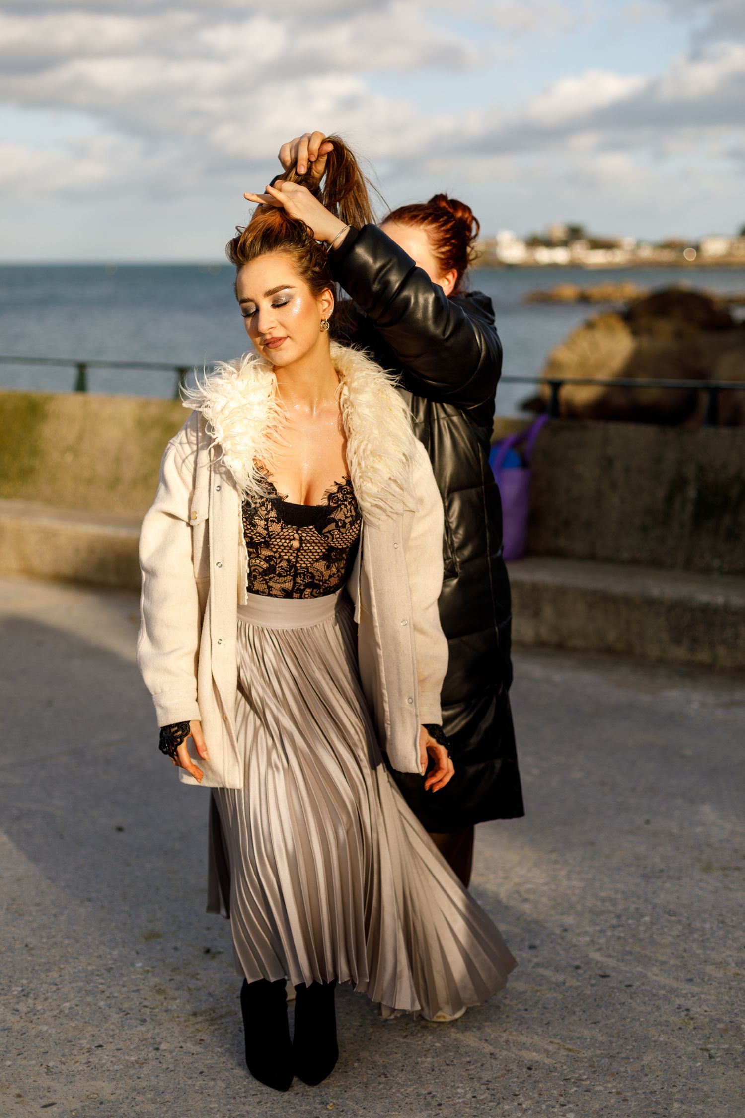 Fashion model in pleated skirt and fur-trimmed jacket poses on coastal walkway during golden hour photoshoot.