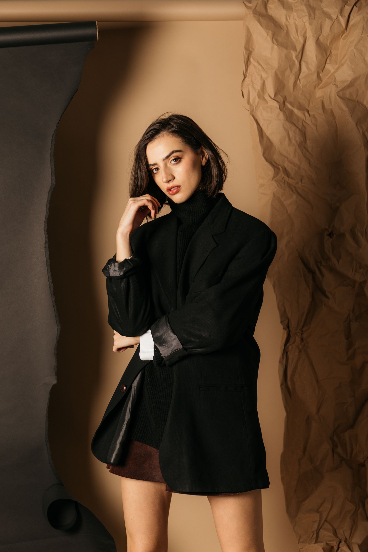 Model poses artistically in black oversized blazer against textured beige backdrop with dramatic lighting.
