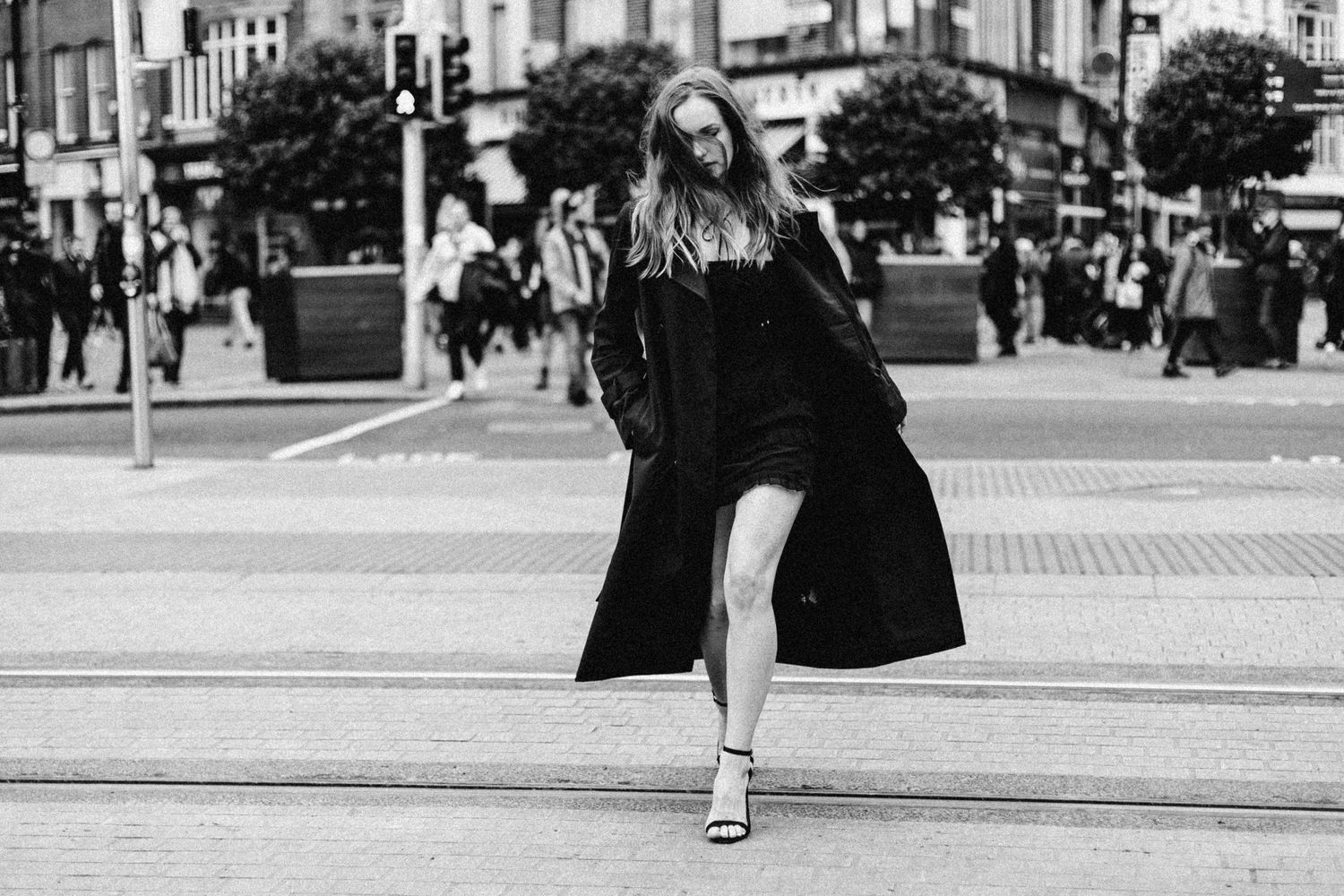 A person in a long black coat dances playfully on a city street in black and white photography.
