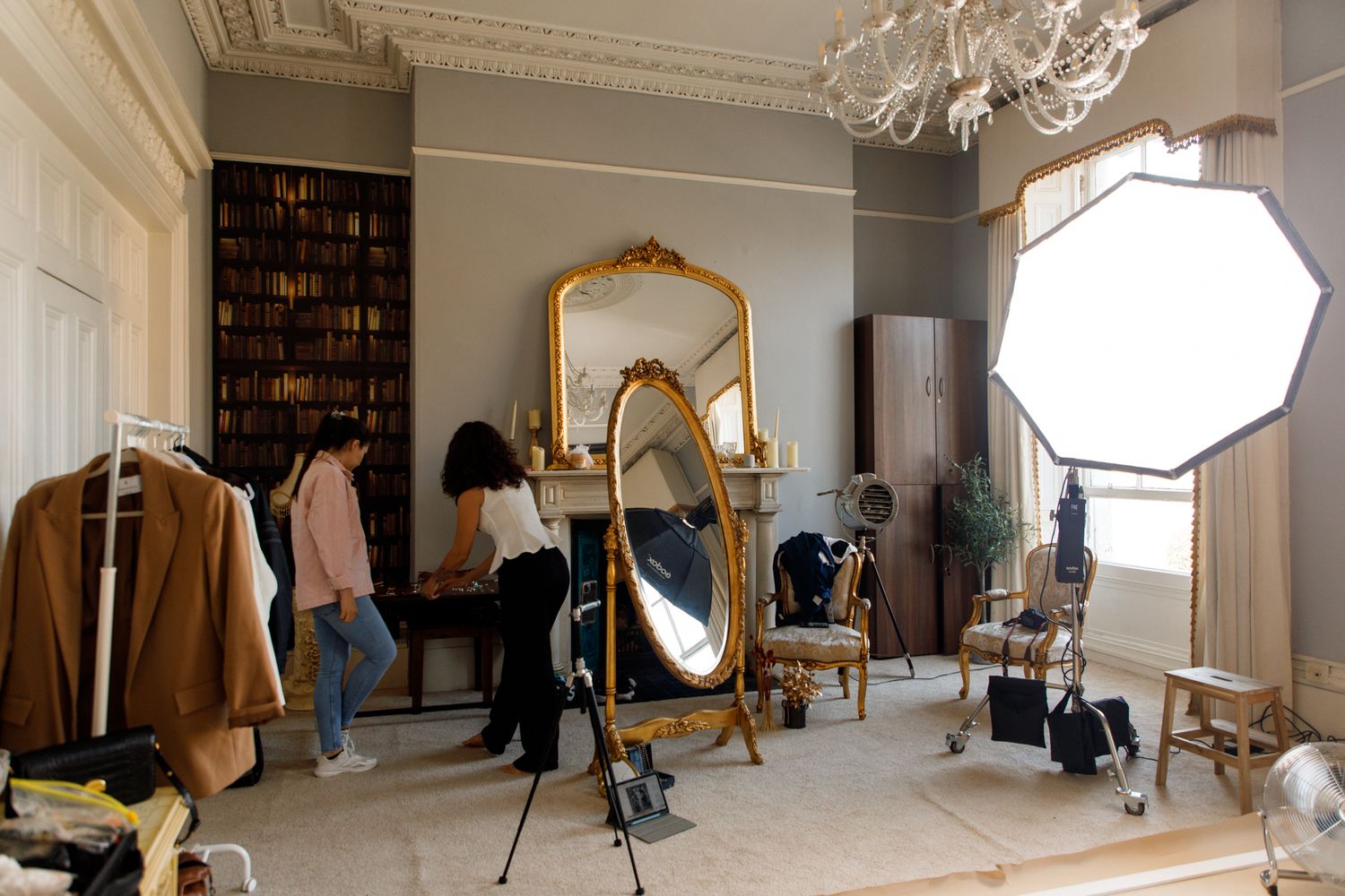 Behind the scenes photo of a fashion photoshoot setup in an elegant room with lighting equipment and ornate gold mirror.