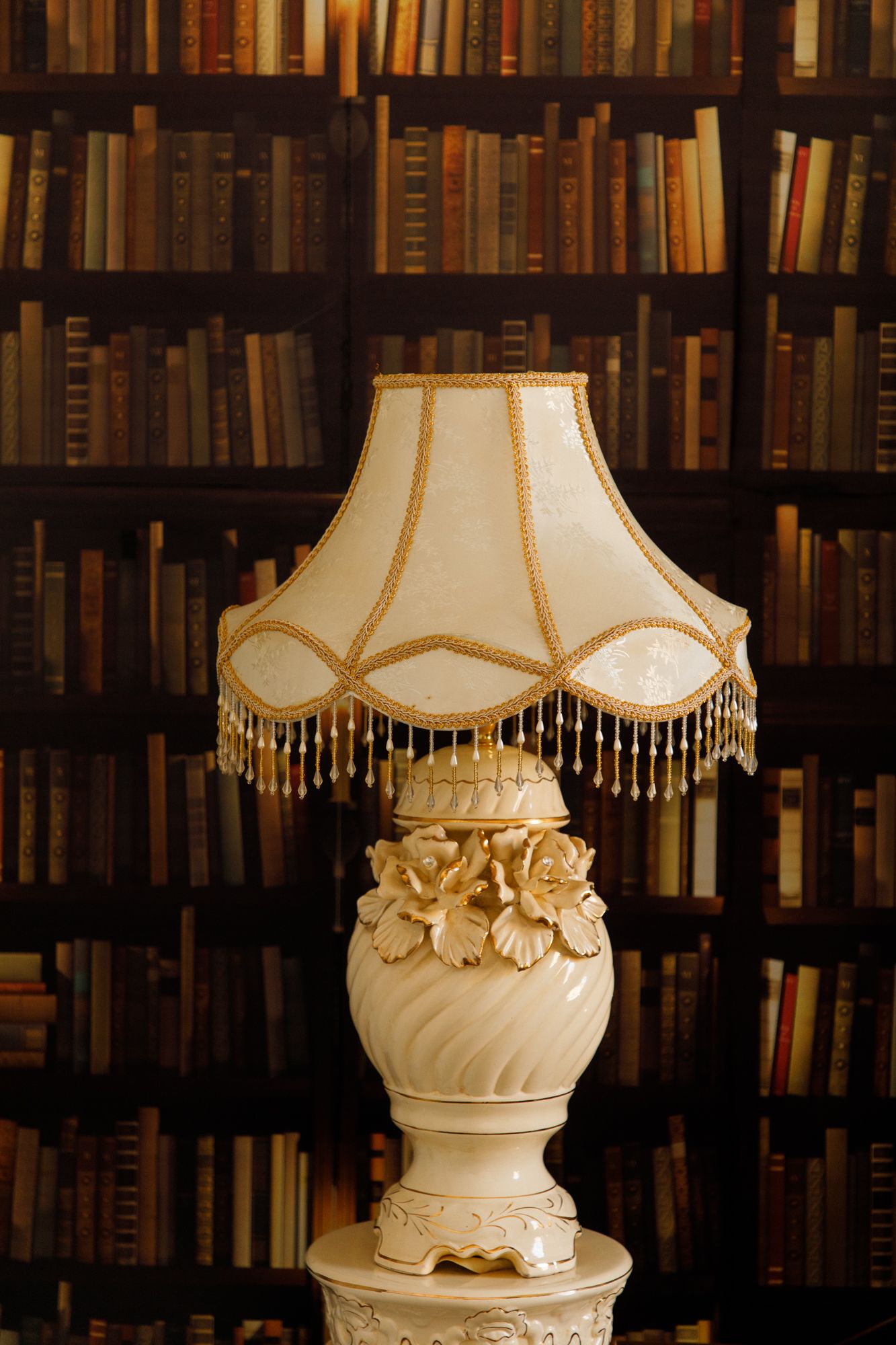 Vintage table lamp with ornate floral base and beaded cream lampshade against bookshelf background in library.