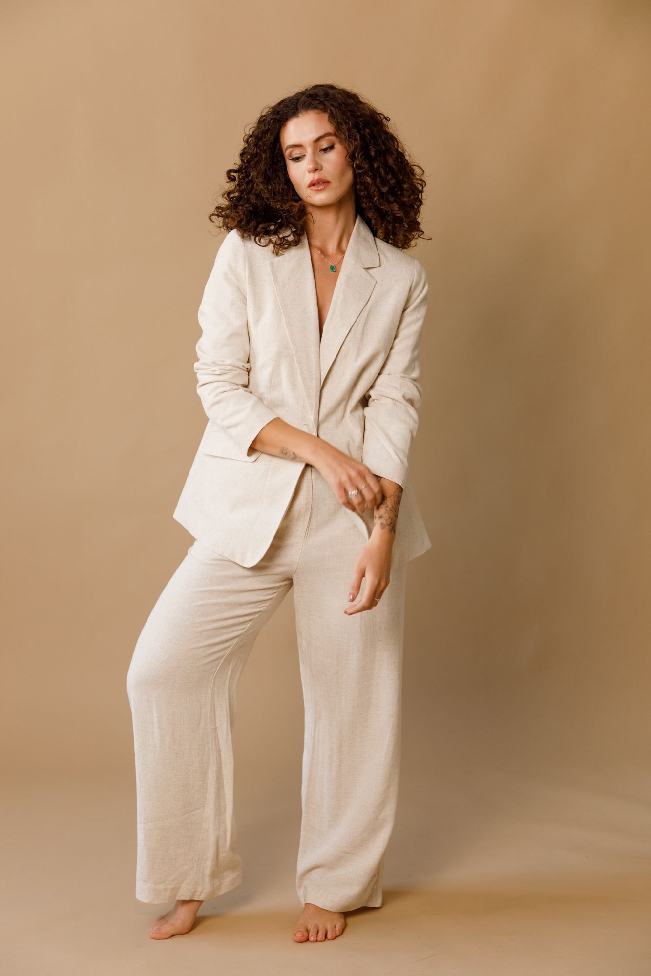 A barefoot model poses in an elegant cream-colored linen pantsuit against a beige background.