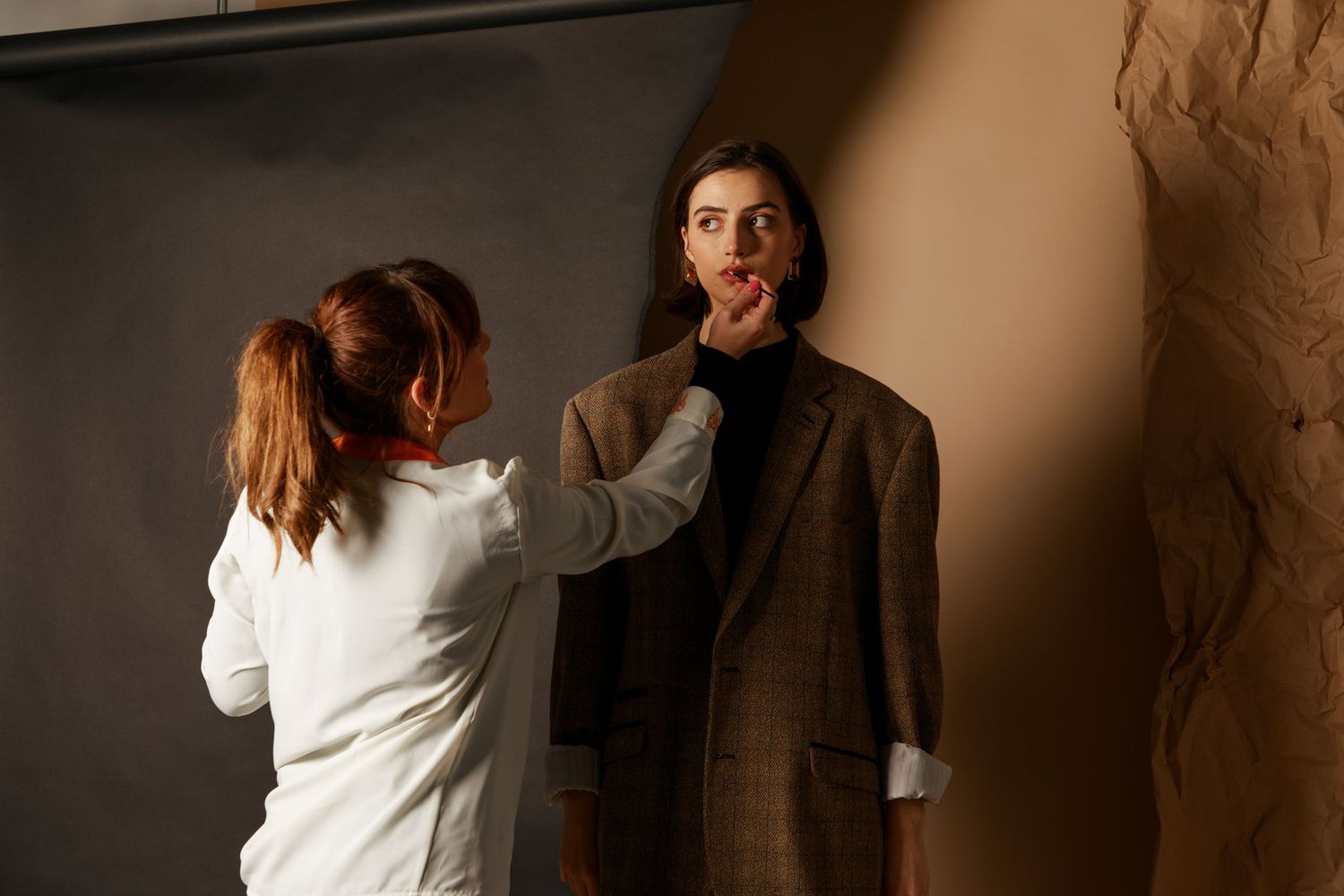 A makeup artist wearing white applies dramatic red lipstick on someone in a brown coat during a photo shoot.