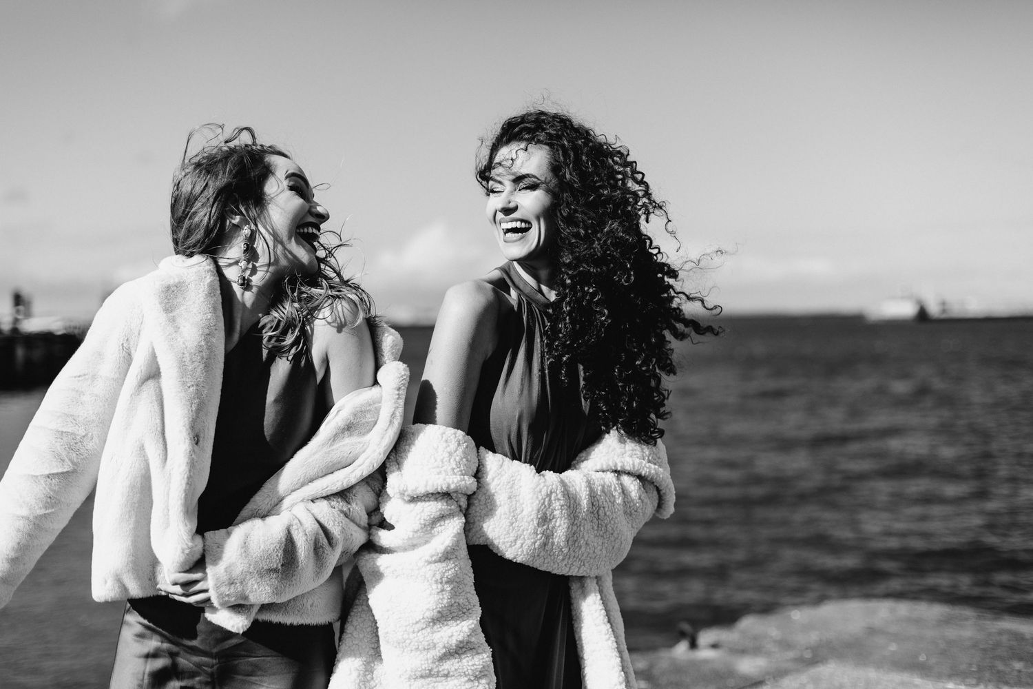 A black and white photo of two people in white sweaters laughing together by the ocean.
