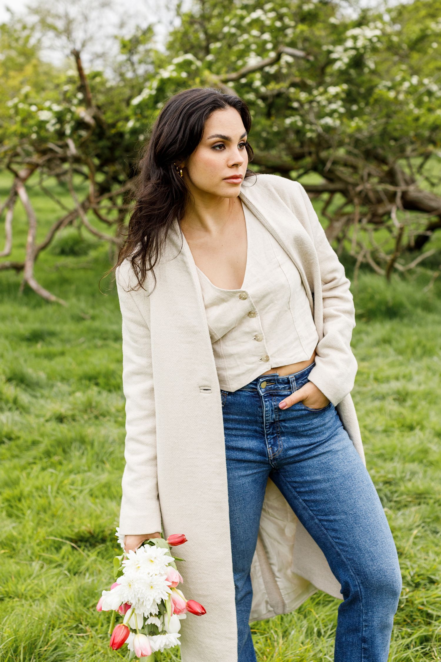A woman in a cream coat and jeans poses with flowers in an orchard setting.