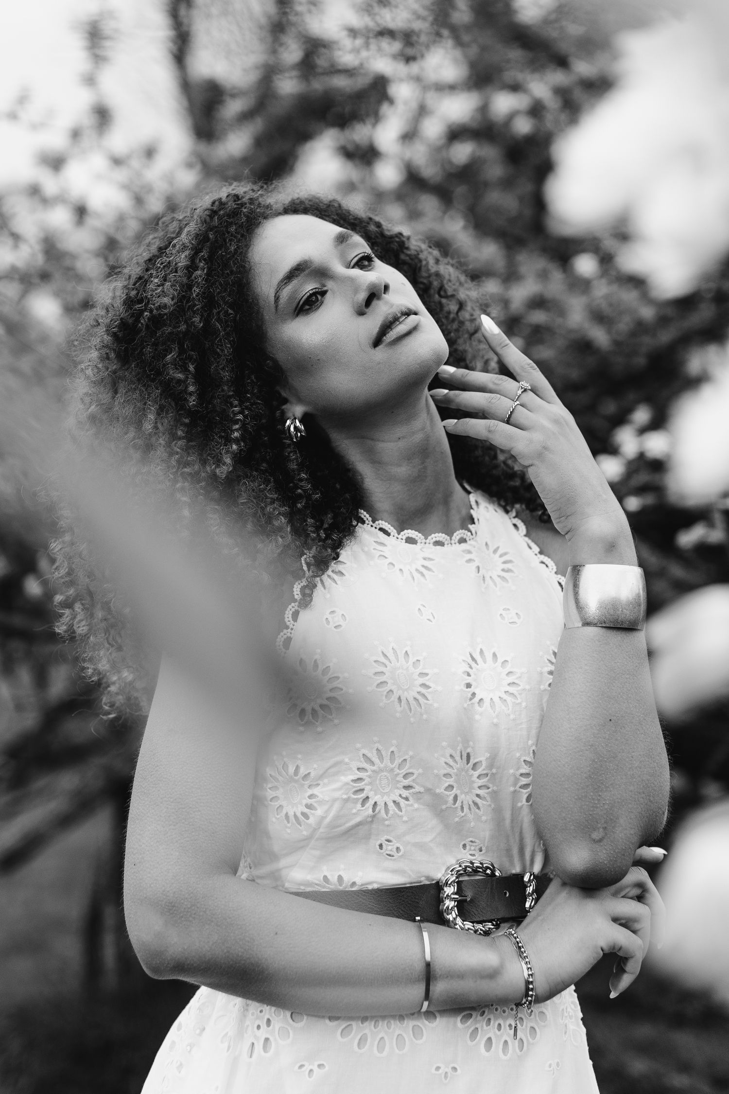 Black and white portrait of a model in white eyelet dress posing outdoors with natural curly hair and dreamy expression.