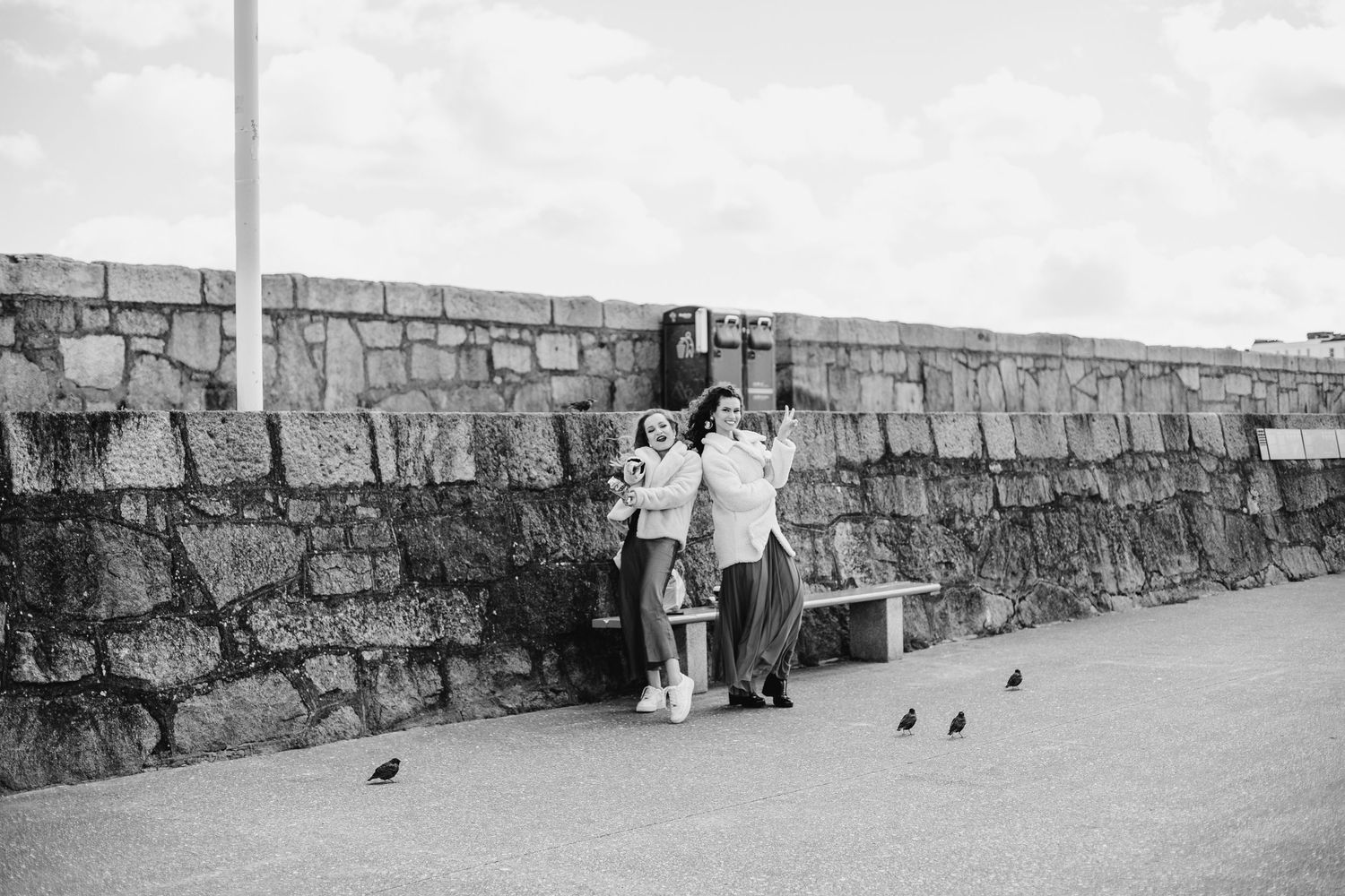 Black and white photo shows three people standing against a stone wall on a promenade with birds nearby.