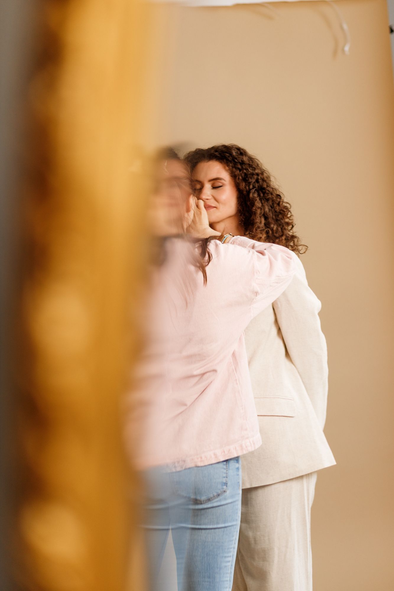Artistic profile portrait through glass showing a person with curly hair in casual clothing against a beige wall.
