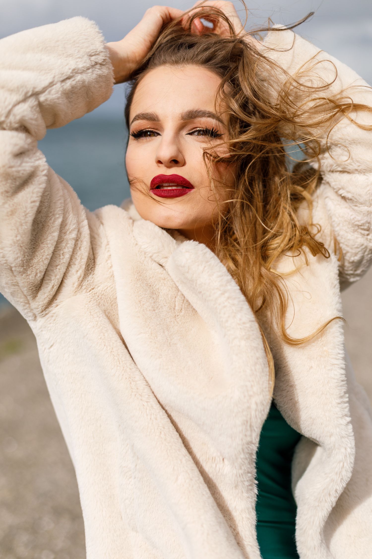 A close-up fashion portrait shows someone wearing a cozy white sweater with wind-blown hair and bold red lipstick.