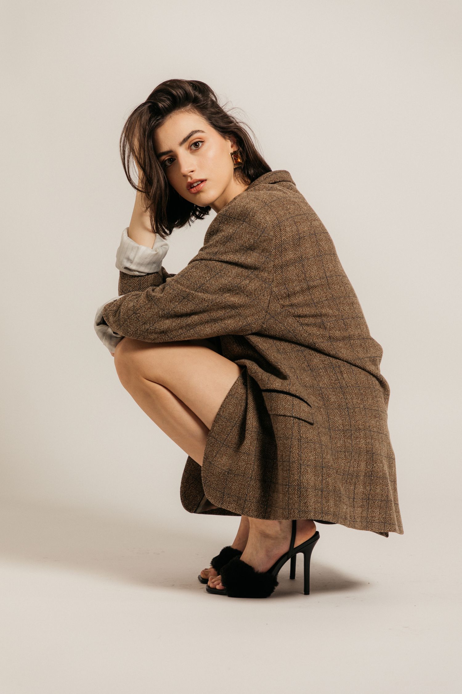 Fashion model poses in brown oversized coat and black heels against neutral backdrop in studio setting.
