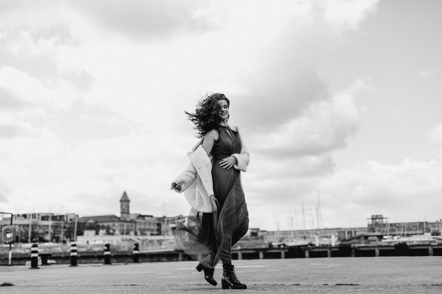A figure dances gracefully in a flowing dress on a pier with a harbor and cityscape in the background.