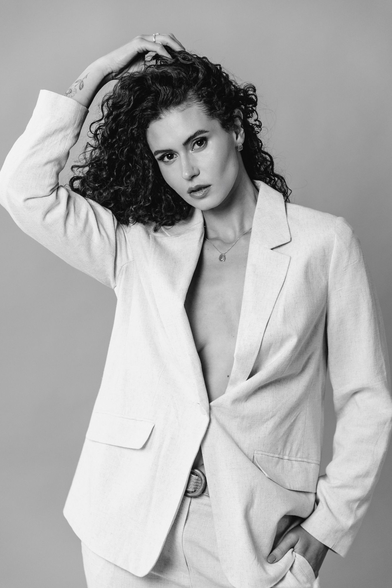 Black and white fashion portrait of a model in a white blazer with curly dark hair against a plain backdrop.