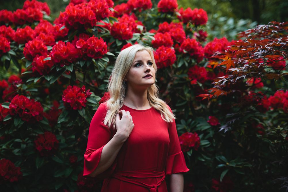 A woman in a red dress poses against vibrant red flowering bushes in a garden setting.