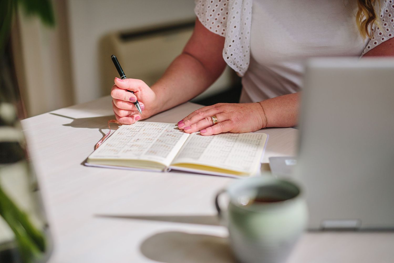 Person writing in journal at white desk with coffee cup and natural light streaming in through window.