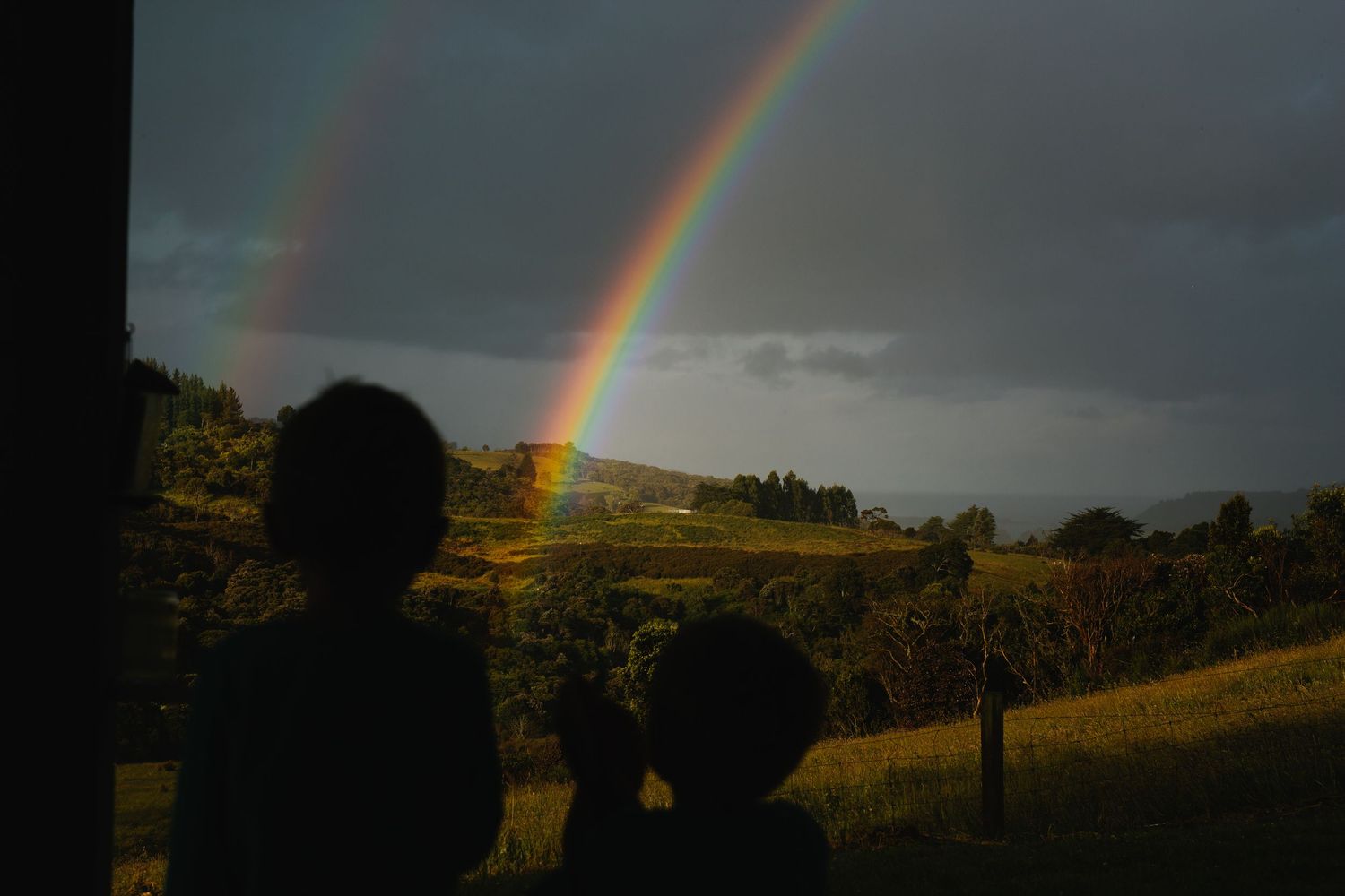 Vibrant double rainbow arches across a moody sky over rolling green hills.