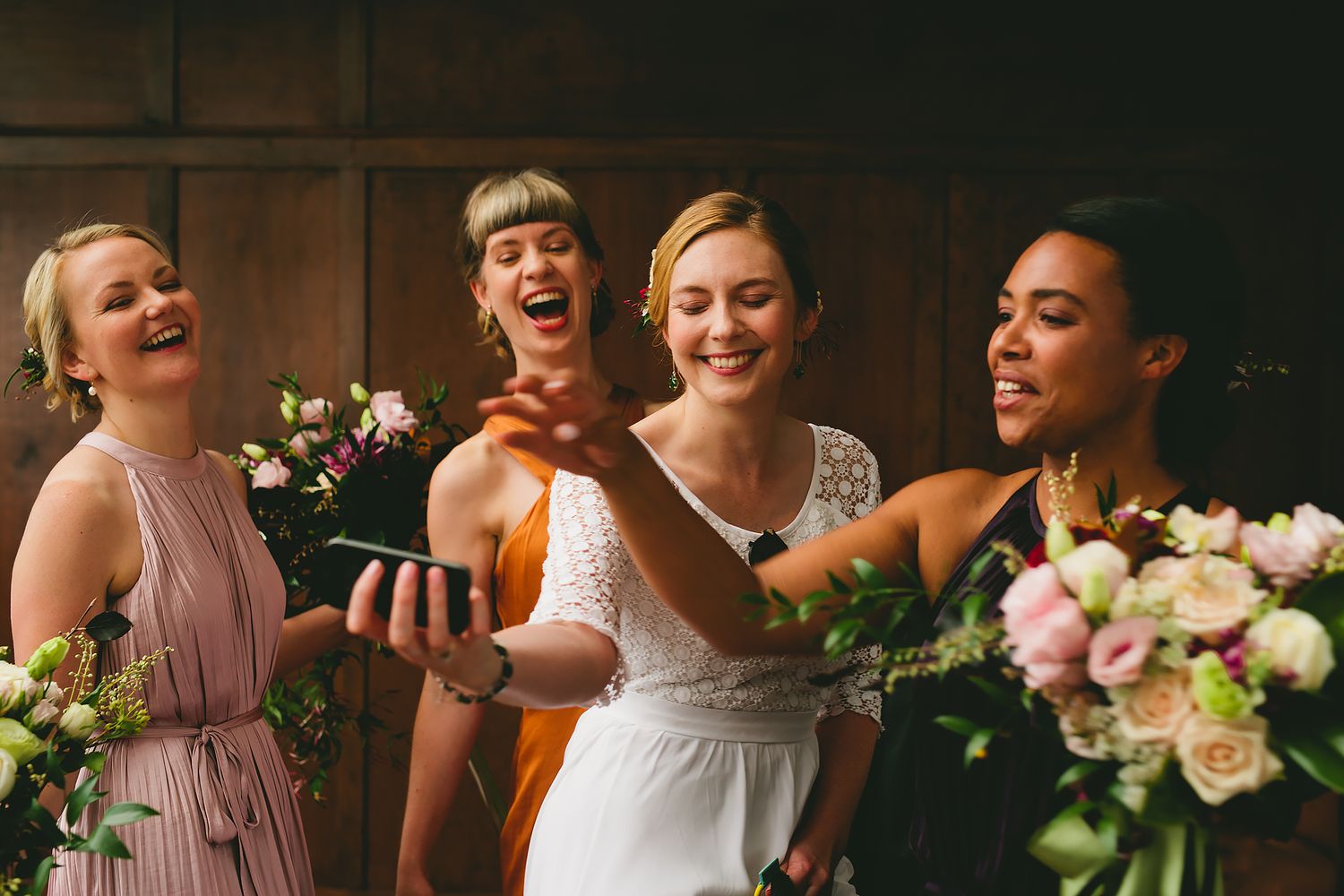 Wedding party members laugh together in a dimly lit venue with floral arrangements.